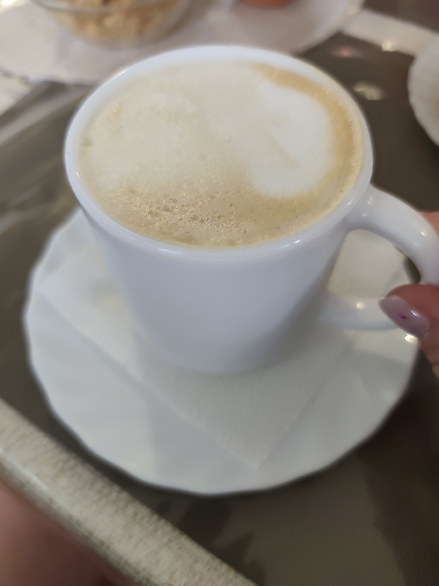 A close-up shot of a white ceramic coffee cup filled with a light-colored coffee beverage topped with foam. The cup is sitting on a white saucer, which is placed on top of a white square napkin. The edge of a table is visible in the bottom left corner of the image, and another dish can be seen partially in the top left corner. A hand with painted nails is visible on the right side of the cup, holding the handle. The background is blurred.
There is no way to know if the image was taken in the city of Нижний Пальник, Russia.