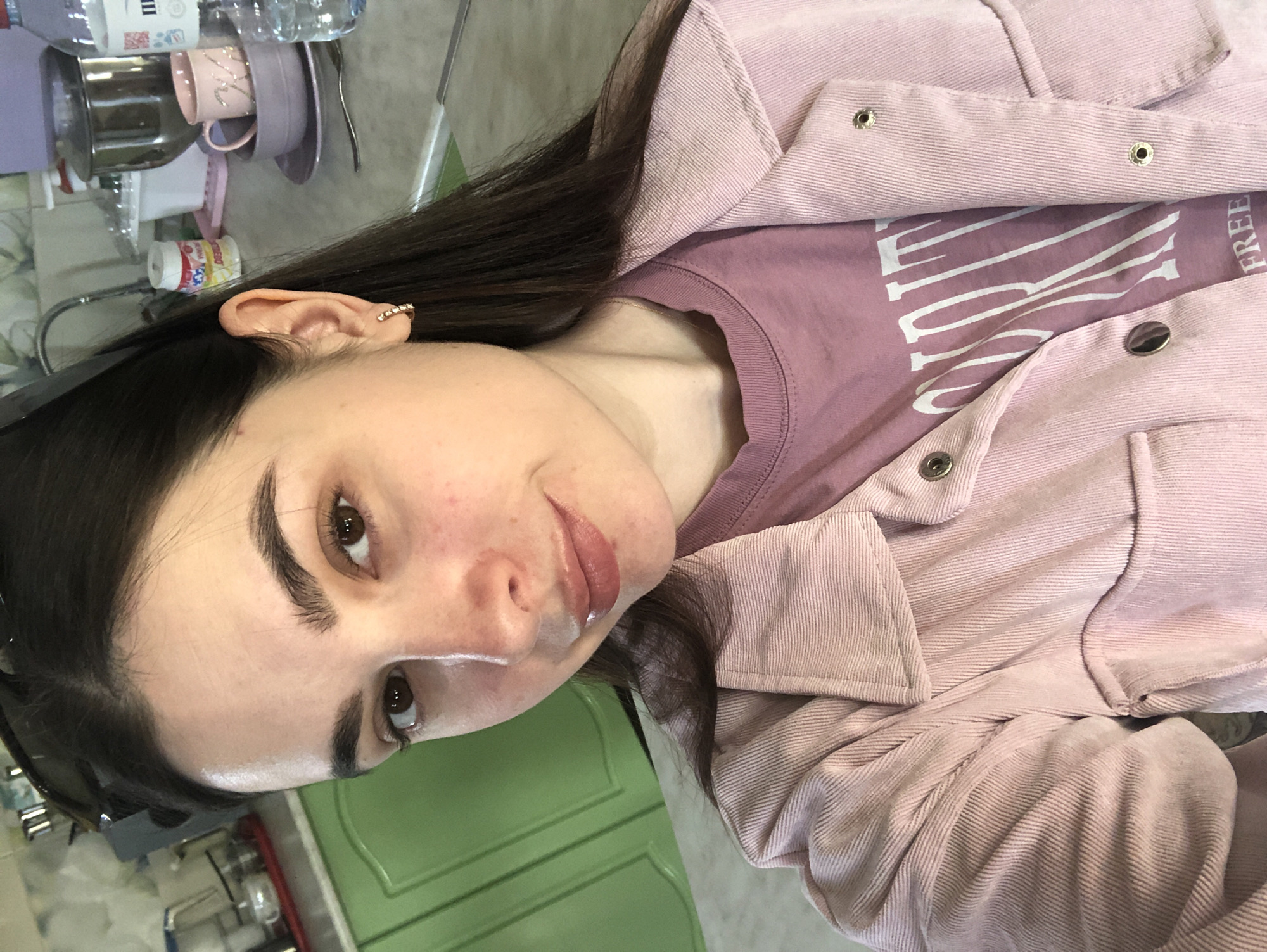 A woman is indoors. She has dark hair, brown eyes, and fair skin. She is wearing a pink corduroy jacket over a purple t-shirt. The room contains a sink, dishes, and a green cabinet. The location is not identifiable from the image provided.