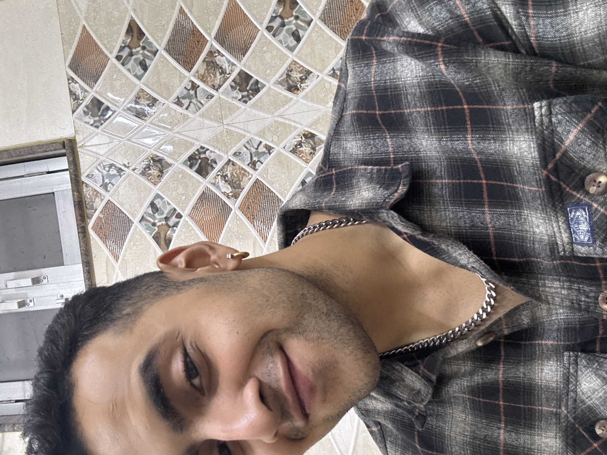 A young man with medium-dark skin, short dark hair, and neatly trimmed stubble looks directly at the camera with a gentle smile. He is wearing a dark gray and black plaid shirt with thin red/orange and white lines, and a silver chain necklace. A gold stud earring with a small white stone is visible in his right ear. A faint scar is present on his forehead.
The man is photographed indoors, likely in Kolkata, India, against a patterned wall. The wall is covered with ceramic tiles arranged in a diamond grid. These tiles feature a mix of beige, various shades of brown, and greenish-gray abstract patterns, with some tiles having a glossy, reflective finish. A metal-framed structure, possibly a window or cabinet, is partially visible on the left side of the frame. The man's shirt also features a brown button and a small, blue, rectangular brand label on the pocket, although the text on the label is not legible. The interior of his shirt's collar appears to be lined with a light brown material. The lighting suggests it is daytime.
