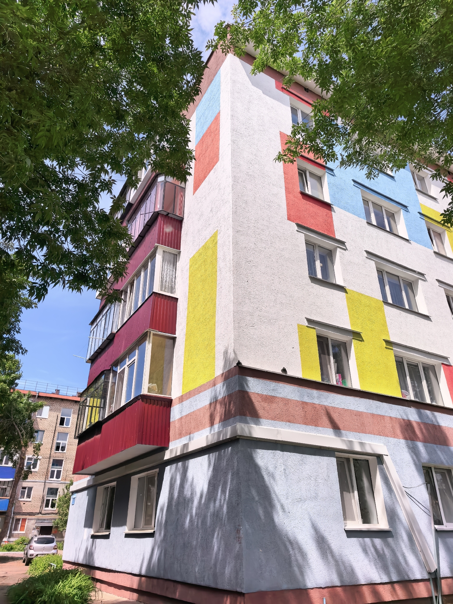 The image shows a corner view of a multi-story building in Sterlitamak, Russia. The building's facade is painted with large blocks of red, blue, and yellow colors on a white background, in a somewhat Mondrian-like style. There are several windows with white frames, some of which have curtains visible inside. A section of the building has balconies enclosed with red corrugated panels and glass windows. The bottom portion of the building is painted with horizontal stripes of blue and red.
The building is partially obscured by green foliage from trees on either side. In the background, another building can be seen. A car is parked on a paved surface near the base of the building. The sky is blue and visible through the trees. The location is Sterlitamak, Russia.