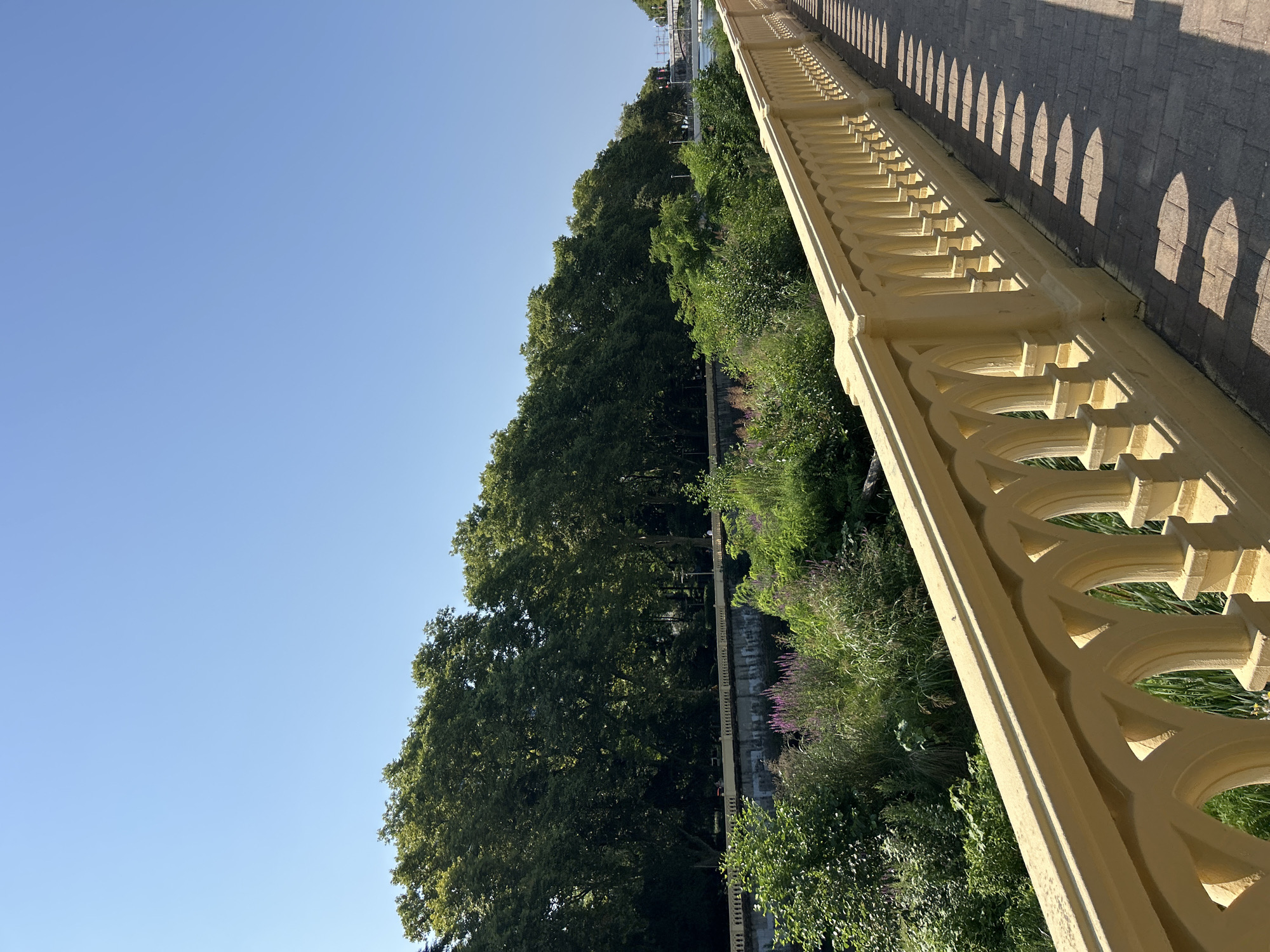 This image captures a scenic view along a tree-lined embankment with a yellow ornate railing in the foreground. The railing runs parallel to a paved walkway, casting distinct arched shadows on the ground due to the bright sunlight. Beyond the railing, lush green vegetation and trees dominate the landscape, with a hint of purple flowers visible in the middle ground. In the distance, a body of water, possibly a river or canal, is visible with a bridge crossing it, and further structures suggest an urban environment. The sky is clear and a vibrant blue. This scene is characteristic of the areas surrounding the Khosta River in Sochi, Russia, where similar promenades and verdant riverbanks can be found. There are no people clearly visible in the frame.