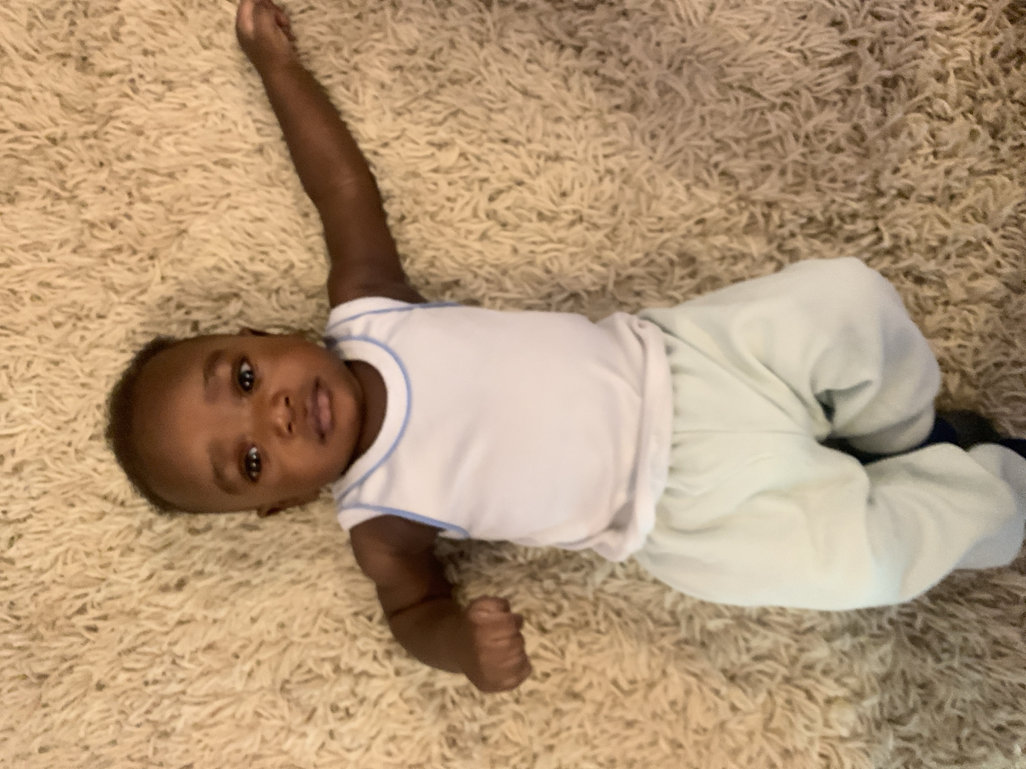 A baby lies on a tan, shag carpet. The baby is wearing a white top with blue trim and light green pants. The baby's left arm is raised. The image was not taken in the city of Barakin Akawo, Nigeria, as that information is unavailable.