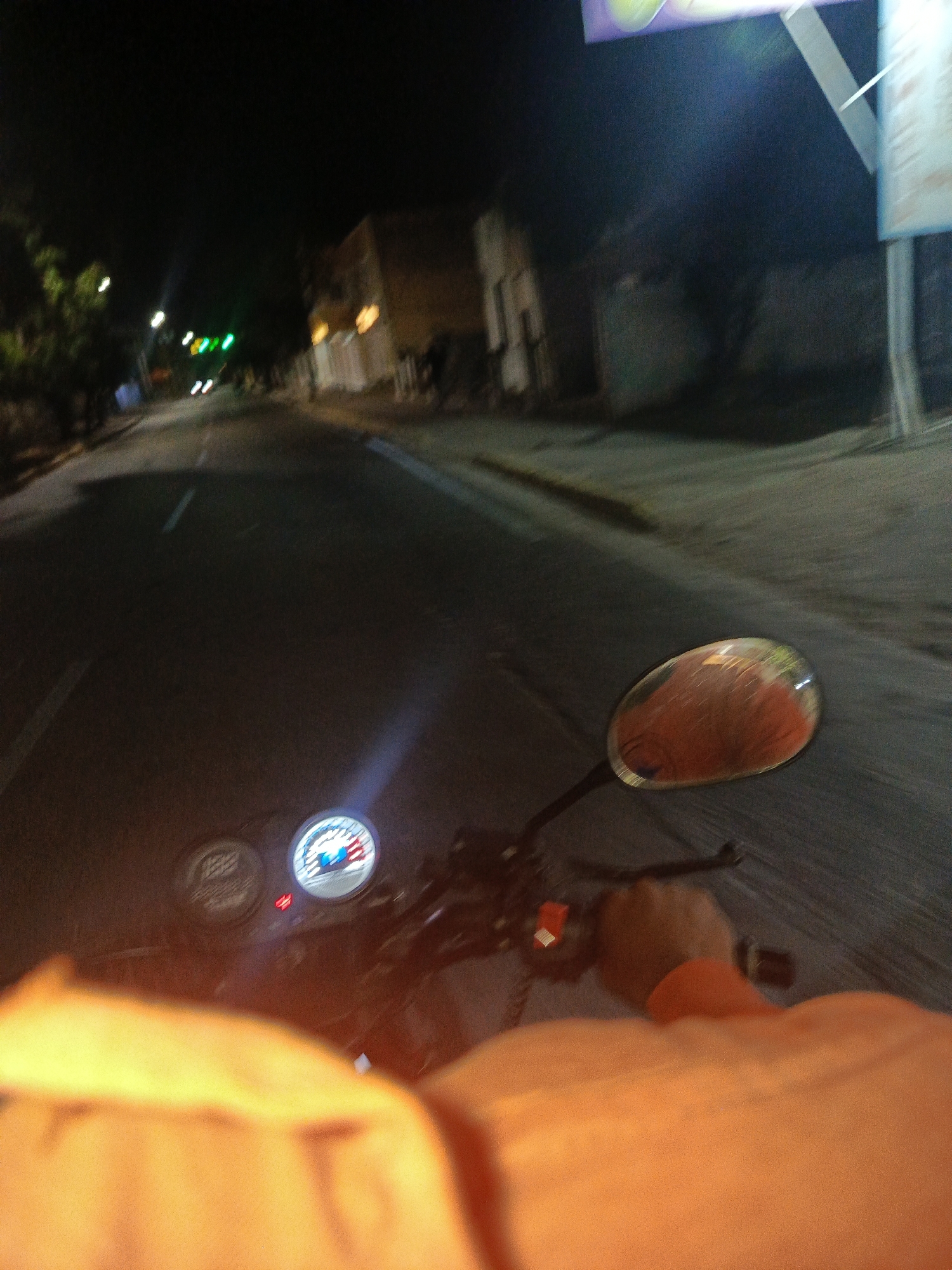 The image is taken from the perspective of someone riding a motorcycle at night in Juan Griego, Venezuela. The motorcycle's speedometer is visible, as is the right side mirror. The road stretches ahead, with buildings and sparse lighting on either side. The street appears to be paved. Trees line parts of the road. The rider's torso and right hand are visible. The rider is wearing an orange shirt or jacket.