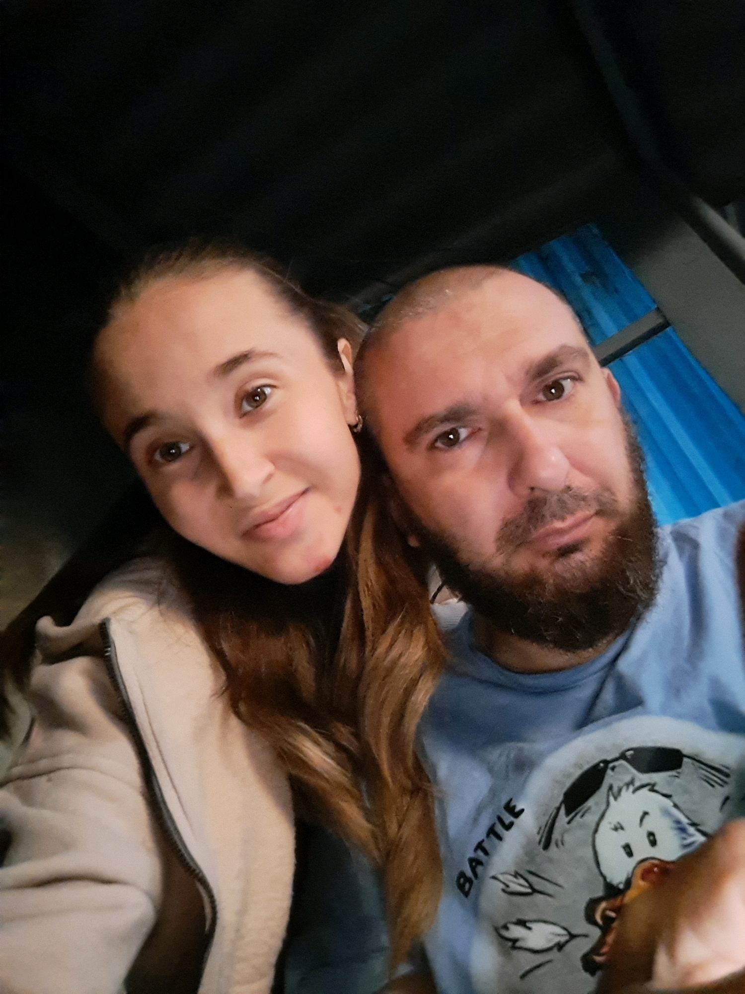 The image is a close-up selfie of two people. On the left is a young woman with long, light brown hair, brown eyes, and fair skin. She is wearing a light-colored zip-up jacket. On the right is a man with a shaved head, brown eyes, and a full beard. He is wearing a light blue t-shirt with a cartoon graphic. They are positioned close together, suggesting an intimate or friendly relationship. The background is dark, with some blue corrugated material visible on the right side. The overall impression is that this is a casual, personal photograph taken indoors or in a sheltered outdoor space. There is no visible information in the image to indicate the location as Vypasne, Ukraine.