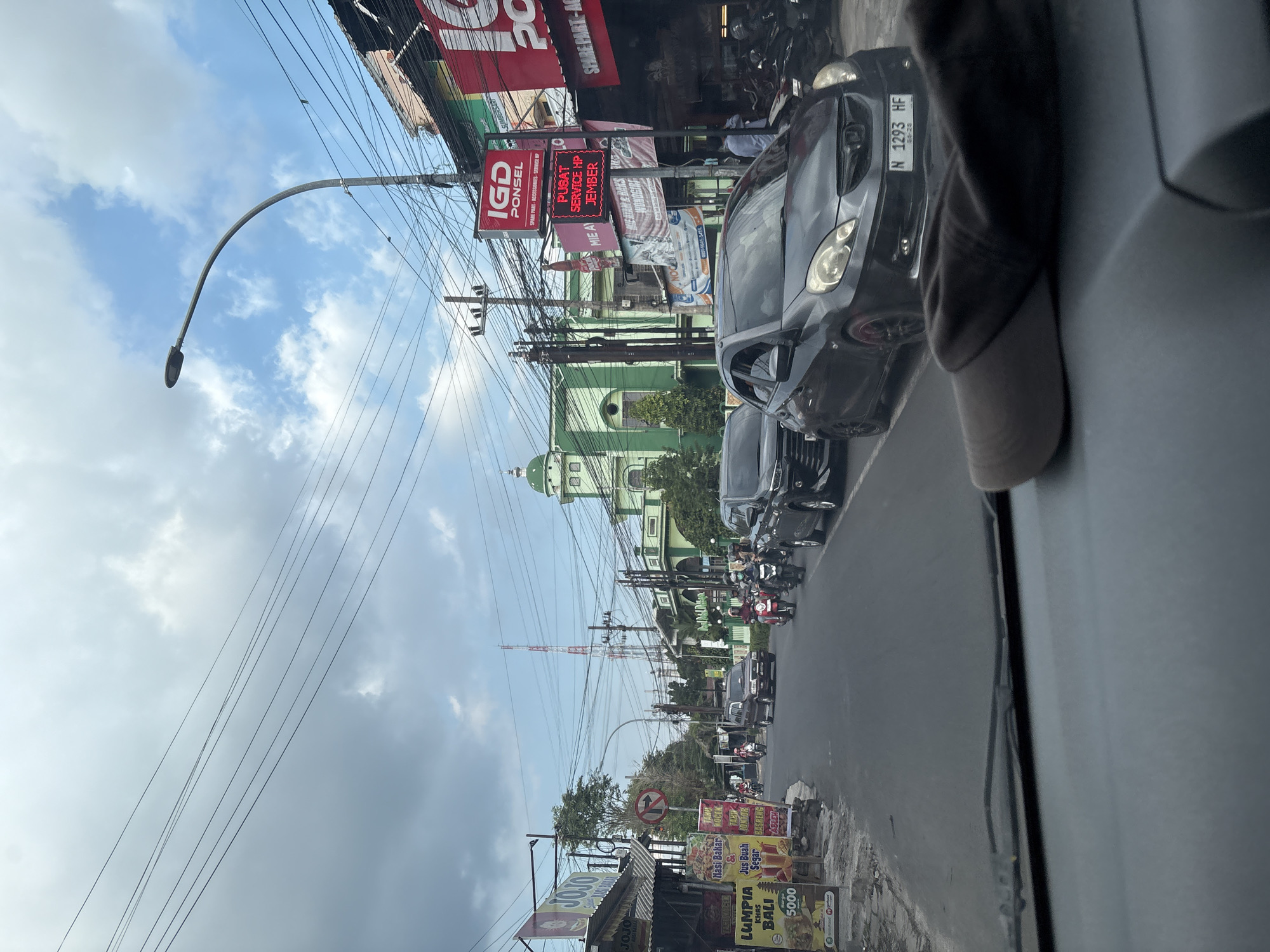 The image shows a street scene in Sumbersari, Indonesia. A gray Honda car with license plate "N 1293 HF" is in the foreground, and a dark baseball cap rests on the dashboard. The street is lined with shops and buildings, including a green mosque with a dome in the background. Several motorcycles and other vehicles are on the road. Overhead power lines and a street light are visible. Signs for "JOJO," "IGD PONSEL," and food stalls are present. The sky is partly cloudy with a blue sky peeking through. The scene appears to be a busy urban environment.