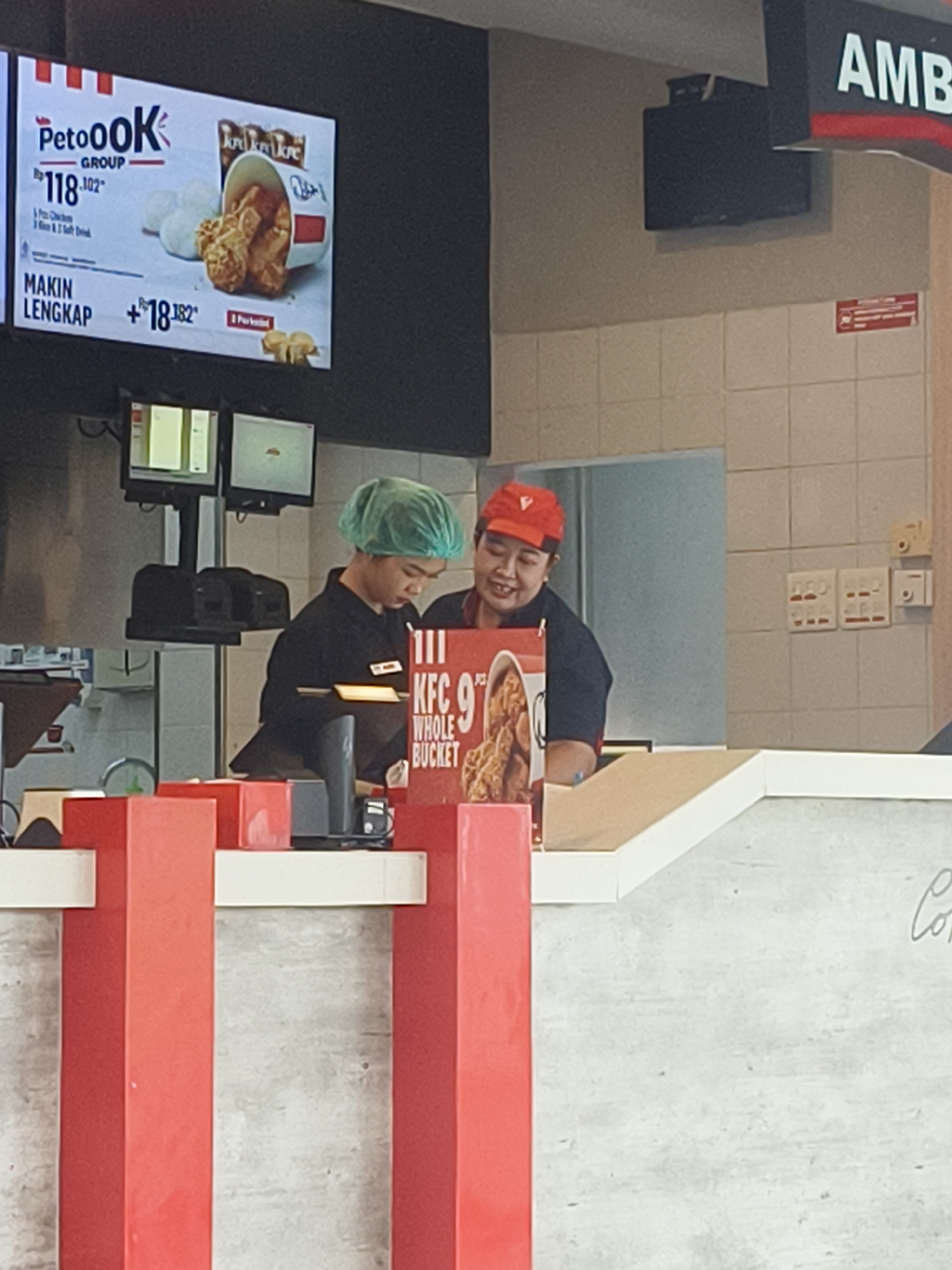 The image shows the interior of a KFC restaurant in Denpasar, Indonesia. Two KFC employees are visible behind the counter. One employee, wearing a green hairnet and a dark shirt, is looking down at something on the counter. The other employee, wearing an orange cap and a dark shirt, is smiling and looking towards the left. A menu board above them displays a "PetoOK Group" promotion with prices in Indonesian Rupiah. A KFC "9 Whole Bucket" display is also visible on the counter. The environment includes a tiled wall, electrical outlets, and bright red structural elements.