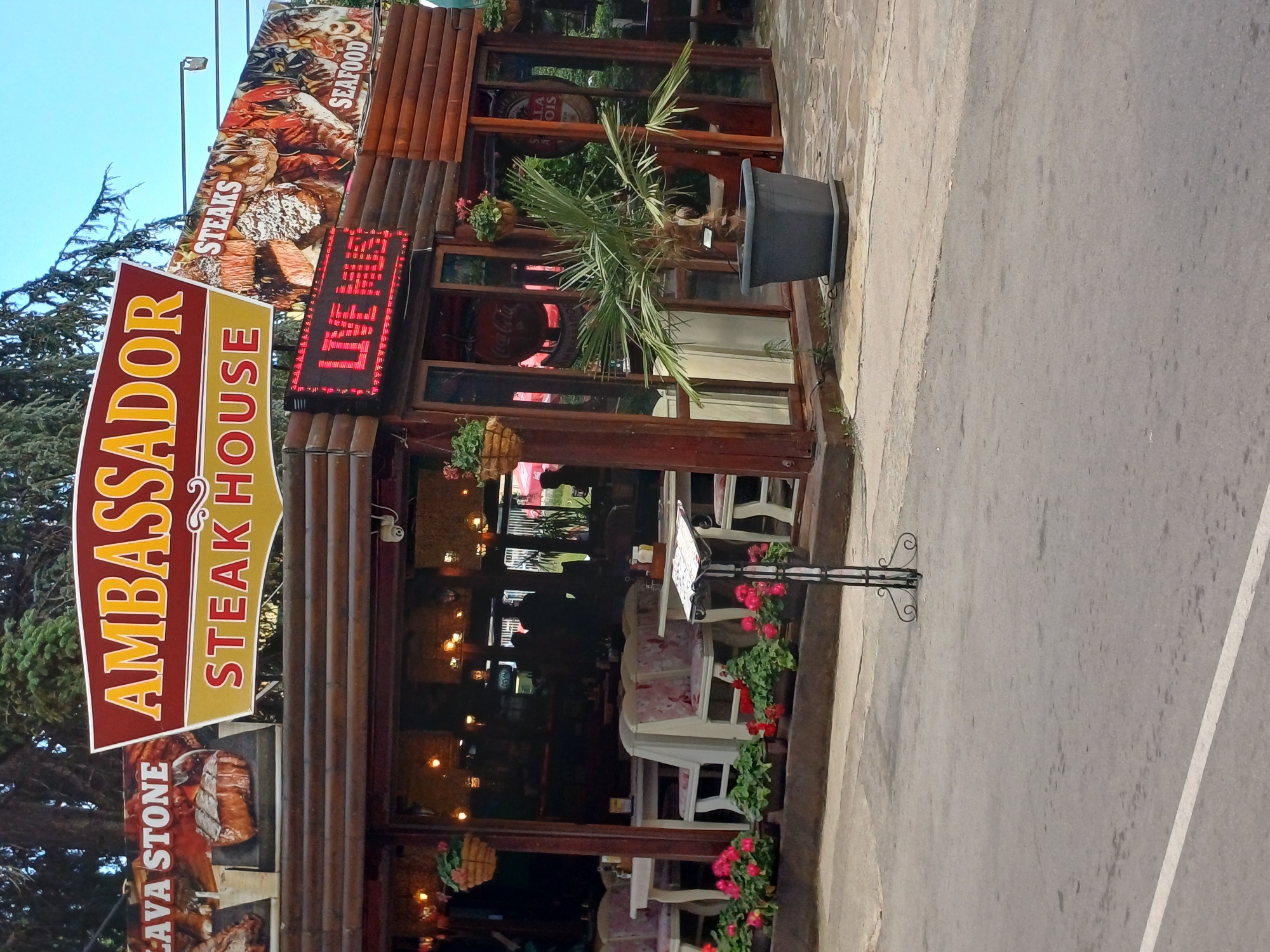 The image shows the exterior of the "Ambassador Steak House" restaurant in Varna, Bulgaria. The building has a rustic, wooden facade with large windows revealing an interior decorated with lights and seating. A prominent red and yellow sign reads "AMBASSADOR STEAK HOUSE." Above the entrance, a large banner advertises "STEAKS" and "SEAFOOD" with images of food. A digital sign displaying "LIVE" in red LEDs is also visible.
The scene appears to be set during daylight, though the lighting within the restaurant suggests it might be approaching evening or is dimly lit. The outdoor area features potted plants and a paved walkway leading to the entrance. A black planter with a small palm tree stands near the building. The overall impression is that of a casual dining establishment. There are no people clearly visible in the image, but the presence of seating and dining elements implies it is open for business. The sky is clear blue.