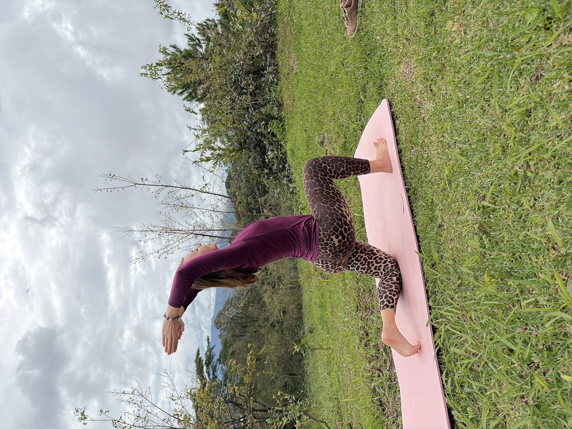 A woman is performing a yoga pose on a pink mat outdoors. She is wearing a purple long-sleeved shirt and leopard-print leggings. Her arms are raised above her head, with her hands clasped. Her left knee is on the mat, and her right leg is bent with her foot on the mat. The background shows a grassy hill with trees and mountains in the distance. The sky is cloudy. A pair of brown shoes are visible on the grass to the right of the mat. This scene is located in Ban Khun Wang, Thailand.