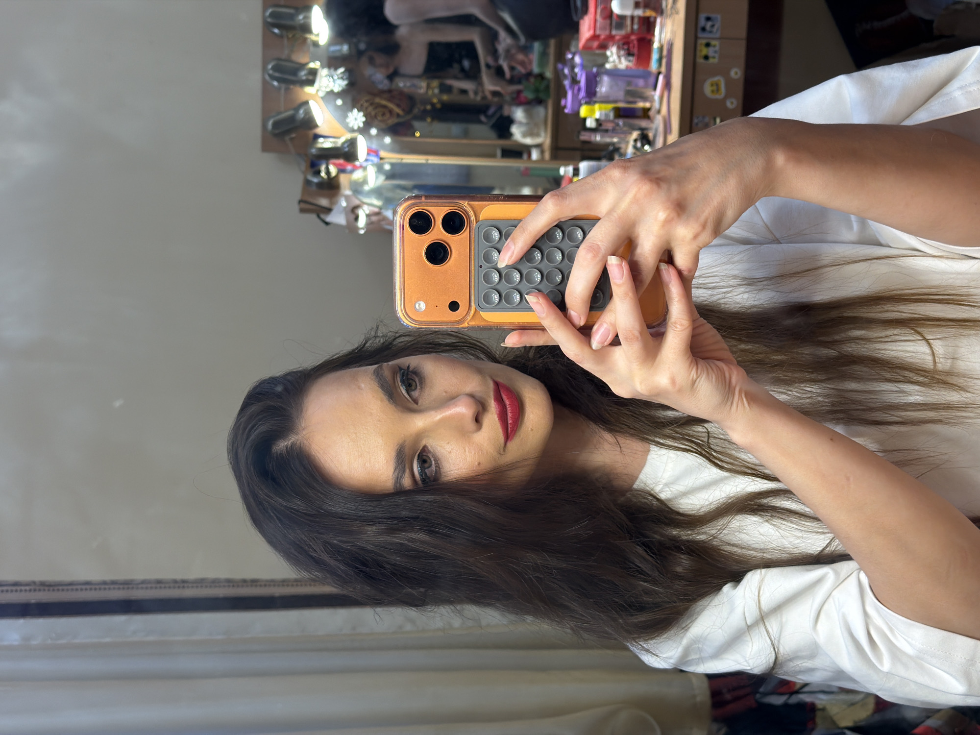 The image shows a young woman with long, dark, wavy hair and dark eyes. She is wearing a white t-shirt and has red lipstick on. She is taking a selfie in a mirror, holding an orange smartphone in her right hand. The phone has a camera array on the back with three lenses and a grey, dimpled grip attached below it.
The background appears to be a dressing area, possibly a vanity or a makeup station. The mirror reflects a cluttered surface with various items, including bottles, containers, and what appear to be makeup or personal care products. Some of the items are illuminated by small lights. In the reflection, a person can be seen behind the woman, partially obscured, suggesting the woman is standing.
The lighting in the image is somewhat dim and diffused, suggesting it might be indoors, possibly in the evening or on a cloudy day. The overall mood of the image is casual and personal, typical of a selfie taken in a private setting. There is no visible text in the image. The location context of Ufa, Russia, does not provide any specific visual cues within the image itself.