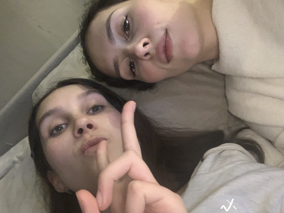 This image depicts two young adult females in an indoor setting. The specific location within Yekaterinburg, Russia, cannot be determined from the visual information provided. The female on the left, positioned closer to the camera, has dark hair, pouting lips, and extends her right hand to form a "peace" sign. She wears a light gray top with a subtle emblem visible on the upper left arm. The female on the right, positioned slightly behind and to the right, has dark, curly hair and looks directly at the viewer with slightly parted lips. She wears a light-colored, possibly cream, sweater. Both individuals appear to be reclining on a soft, light-colored surface, consistent with a bed or sofa, under ambient indoor lighting conditions.