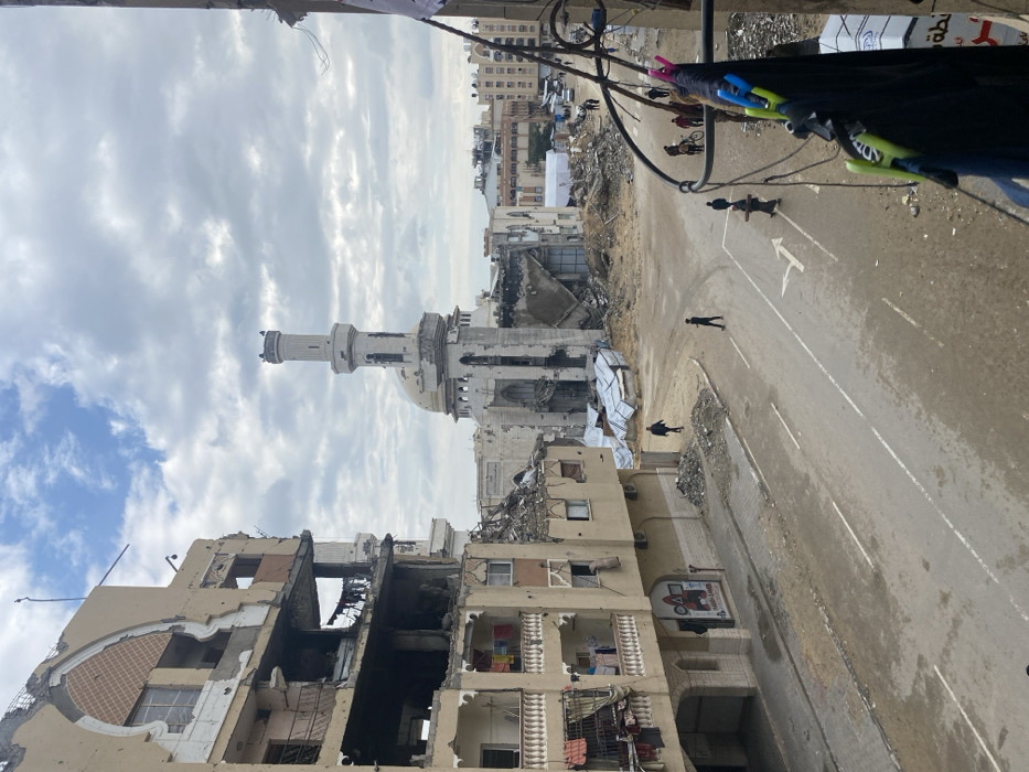 This image depicts a scene of extensive urban destruction in Khan Yunis, Palestinian Territory, focusing on the vicinity of the Great Mosque of Khan Yunis (Al-Omari Mosque).
In the immediate foreground, to the right, a section of a balcony railing is visible with dark clothing items secured by blue, pink, and green clothespins, suggesting habitation or use of the vantage point for the photograph.
To the left, a multi-story residential building exhibits severe structural damage, particularly on its upper floors where sections of walls are missing, and interiors are exposed. Lower floors show partial damage, with clothes visible hanging on several balconies. Rubble is piled at the base of this building and a storefront sign with Arabic text is visible on the ground floor.
The midground features an asphalt road with faded white double-lane markings and a right-turn arrow painted on the surface. The road appears dirty and potentially wet. Several individuals are present on the street: two men walk on the left side, one in dark clothing and another in lighter attire; further down, a small group of people, including one with a bicycle, are moving; another solitary figure walks near the center of the intersection. The area surrounding the road consists of vacant lots filled with extensive piles of concrete rubble, dirt, and twisted sheets of corrugated metal.
Dominating the background is the severely damaged Great Mosque of Khan Yunis. Its prominent minaret remains mostly standing but shows signs of impact. The main prayer hall structure beneath the mosque's dome is largely destroyed, with significant portions of its walls collapsed and its interior exposed to the elements. Blackened areas on the mosque structure suggest fire damage. Beyond the mosque, additional damaged multi-story buildings and a white tent-like structure are discernible. The sky is overcast with thick, grey clouds.