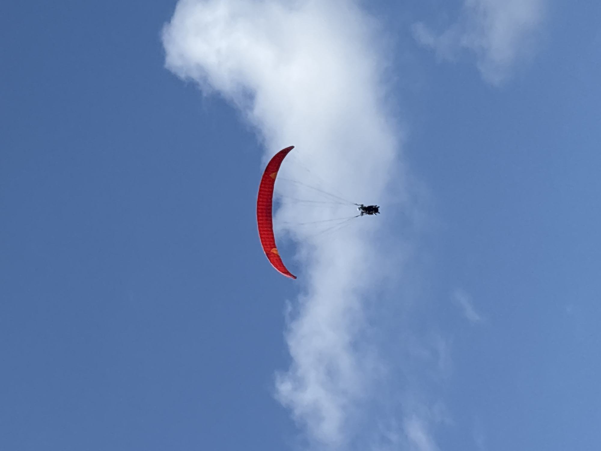 A single person paraglides under a vibrant red canopy against a clear blue sky with scattered white clouds. The paraglider is suspended in the air, appearing to be in motion. The image captures the vastness of the sky and the isolation of the paraglider, suggesting an outdoor recreational activity taking place at a considerable altitude. The scene is devoid of any urban or natural landscape, focusing solely on the paraglider and the sky.