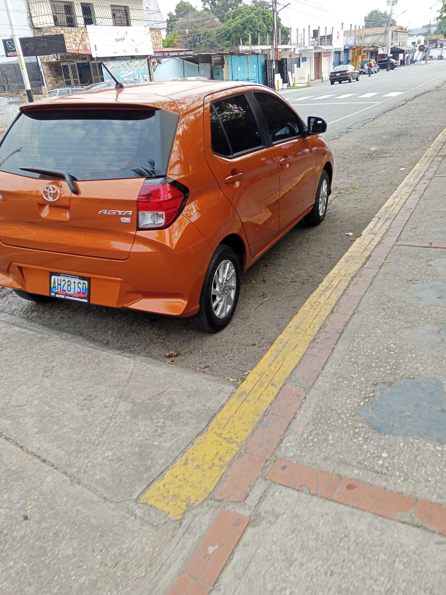 An orange Toyota Agya hatchback, bearing Venezuelan license plate AH281SD, is parked or stopped parallel to the curb on the right side of a two-lane asphalt road in Tinaquillo, Venezuela. The vehicle's rear is prominently featured, displaying tinted windows and a roof-mounted antenna. The immediate foreground shows a light gray concrete sidewalk with a yellow-painted stripe and a section of red brick pavers bordering the asphalt. The asphalt road exhibits minor debris and stains, with white pedestrian crossing stripes visible further down. The left side of the road is lined with various commercial buildings. One notable structure features a stone facade on its upper level with barred windows, and multiple shopfronts below, some with metal security shutters. Visible signage includes "Barquilla" and "NESCA," along with a partially visible sign listing vehicle brands such as "CHEVROLET FIAT DAEWOO CHERY HYUNDAI." Several other vehicles are visible in the distance on the road, including a dark sedan and a white van, with one individual appearing to stand near the dark sedan. Electrical poles and overhead wires are present, and green foliage from trees is visible in the background, suggesting an urban environment with vegetation under daylight conditions. No immediate actions or interactions involving people are discernible in the immediate foreground.