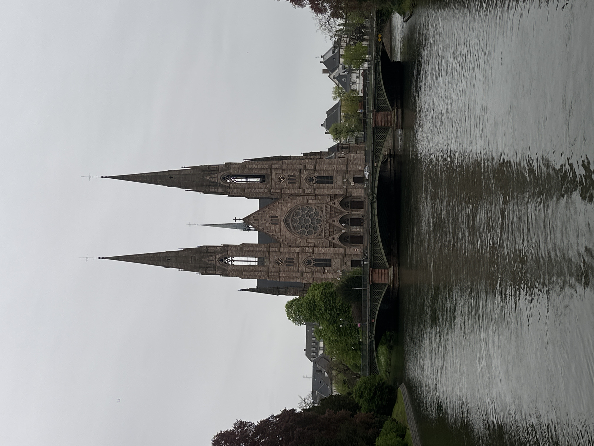 The image depicts the Saint Paul's Church in Strasbourg, France, viewed from across the Ill River on a cloudy day. The Gothic Revival church features two prominent spires and a rose window. A bridge spans the river in front of the church. Trees and buildings are visible along the riverbanks. The water reflects the sky and surrounding architecture.