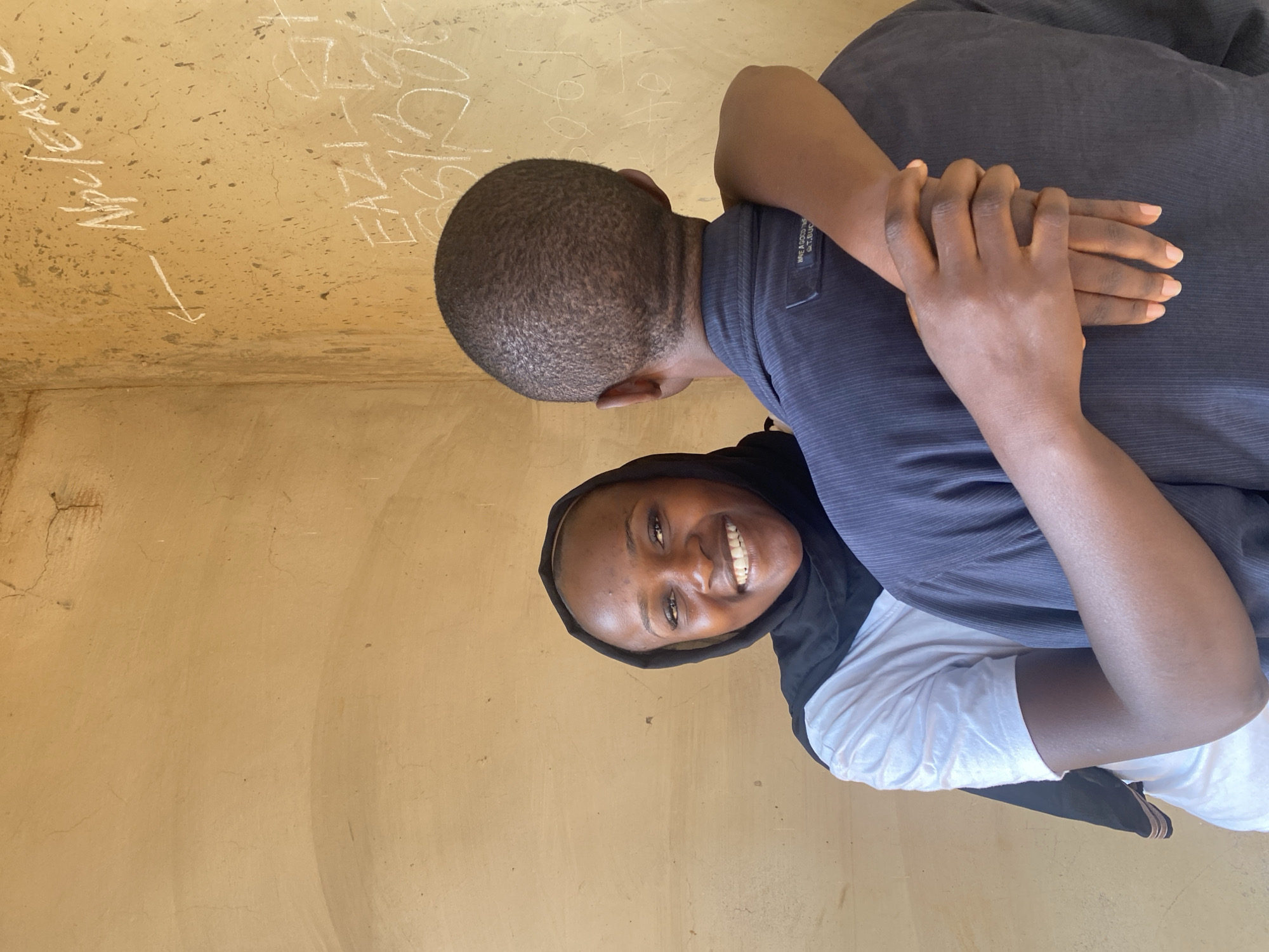 The image features two individuals embracing in what appears to be an indoor setting with textured, light brown walls. The photo is taken from a slightly high angle and rotated, showing the subjects sideways.
One person is seen from the back, with their head turned away from the camera. They have short, dark hair and are wearing a dark grey or black collared shirt. Their arms are wrapped around the second person. A small, unreadable label is visible on the shirt's upper left side.
The second person is facing the camera, smiling broadly and looking directly at the viewer. They are wearing a black head covering, possibly a hijab or headscarf, and a white shirt. Their expression is joyful and engaging, with a clear, wide smile.
The background wall is a prominent feature, displaying a rough, possibly plastered texture with some cracks and speckles. Several inscriptions in white chalk-like material are visible on the wall. On the left, "Npc/EAP" is clearly legible, with an arrow pointing downwards and to the right. Further to the right, the letters "EAZ" can be seen, followed by other less distinct characters, possibly including "SYZ" or "S/L". The lighting is bright and even, suggesting it is daytime. The overall scene conveys a sense of warmth and happiness, set in Kuru Dungus, Nigeria.