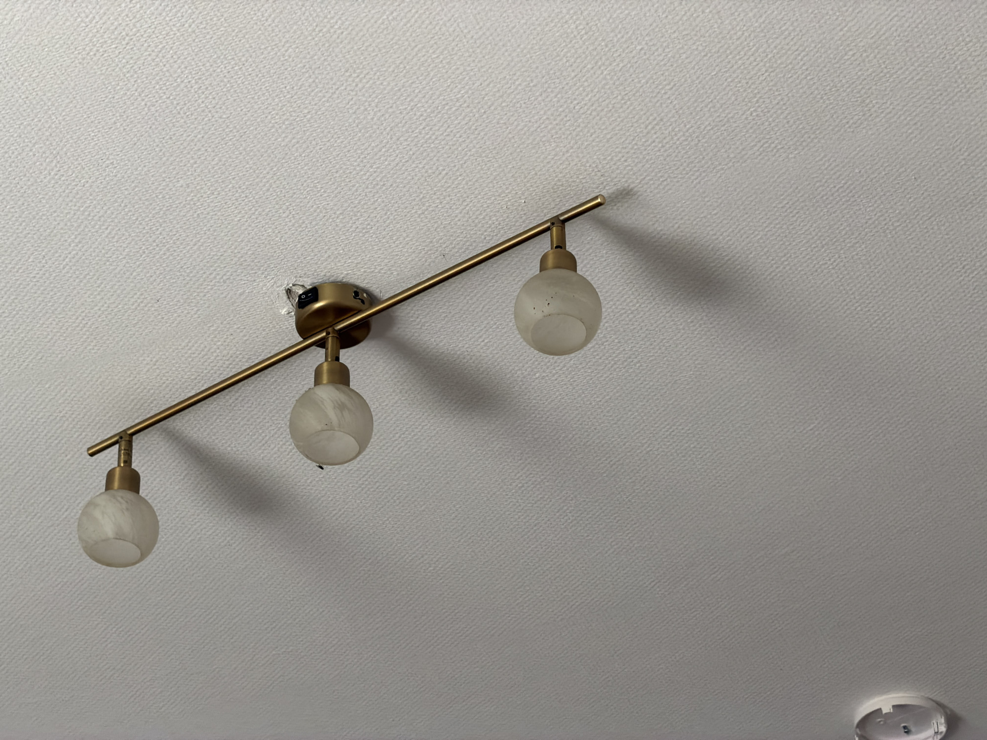 A brass track light fixture with three frosted glass globe lights is mounted on a textured white ceiling. The track runs horizontally across the frame, and the lights are suspended from it. A small black switch is visible on the base of the fixture where it attaches to the ceiling. The ceiling has a fine, uniform texture. There is a shadow cast on the ceiling by the light fixture. In the lower right corner of the image, a small portion of a white circular object, possibly a smoke detector, is visible. The scene is indoors.