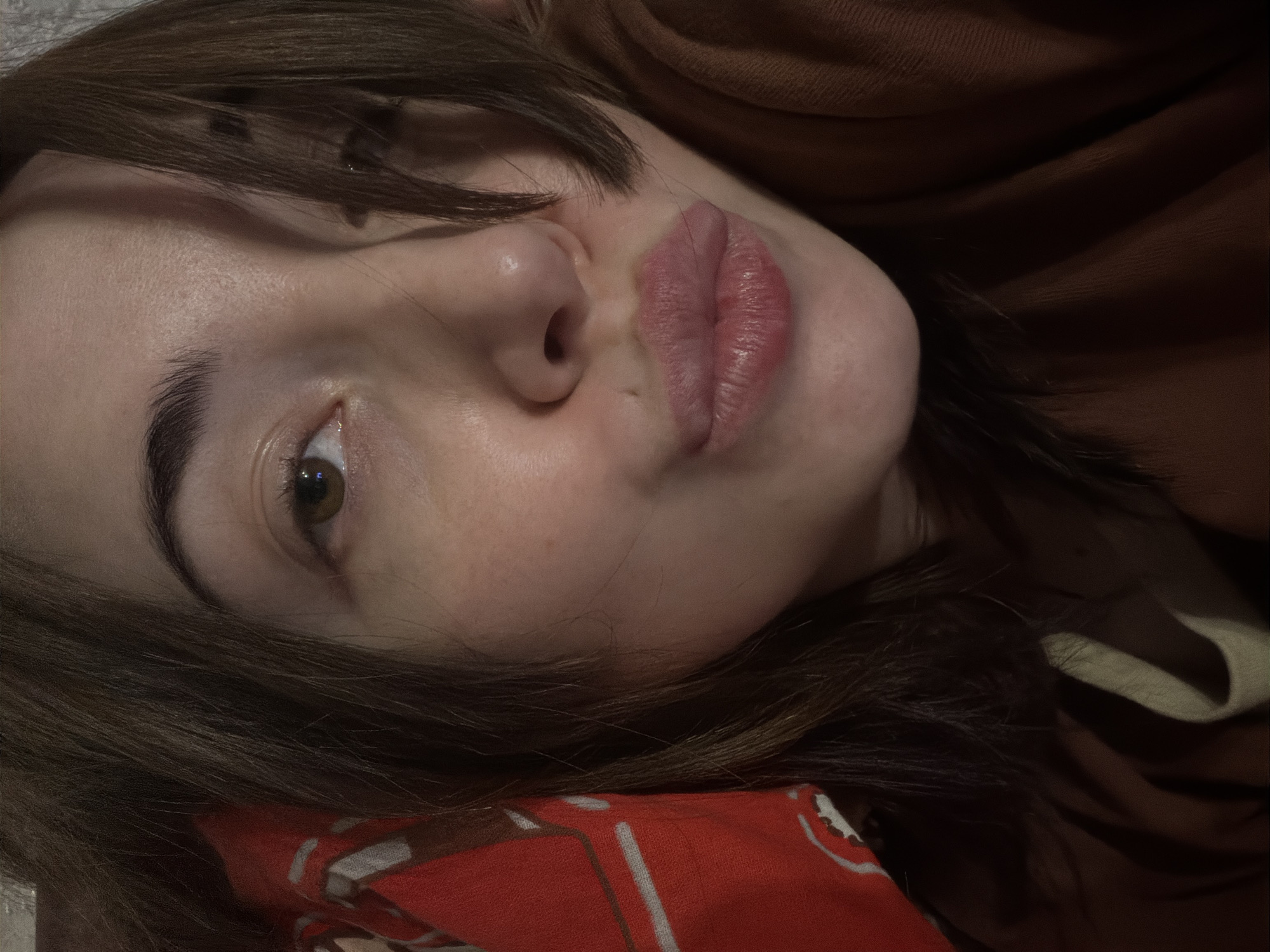 A close-up, head-and-shoulders shot captures a young woman with medium brown hair, her right eye visible, and her lips pursed in a kiss. She is wearing a brown sweatshirt, and a red and white patterned fabric is visible behind her left shoulder. The lighting is dim, casting soft shadows on her face. The background is indistinct.