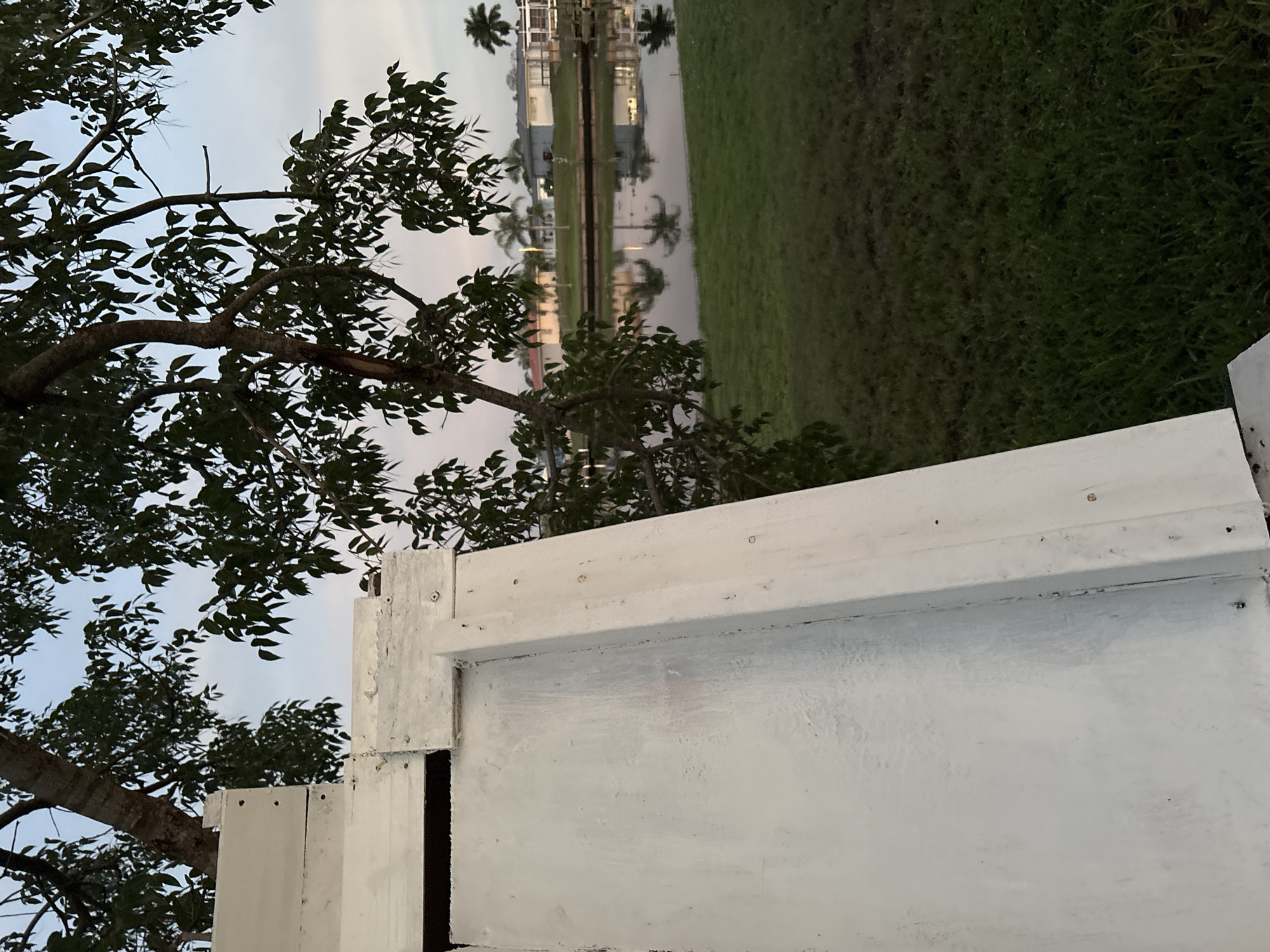 The image shows a close-up view of a white wooden structure in the foreground, possibly part of a building or a fence, with a tree and its branches extending into the frame from the left and top. Beyond the structure and tree, a body of water, likely a canal, is visible, reflecting the sky and palm trees. On the far side of the water, residential buildings with light-colored walls and roofs can be seen, typical of a suburban or coastal setting. The sky is overcast with a hint of pink and blue, suggesting it is either sunrise or sunset. The overall environment appears to be a residential area in North Port, Florida, characterized by its canals and tropical vegetation. No people or animals are present in the image.