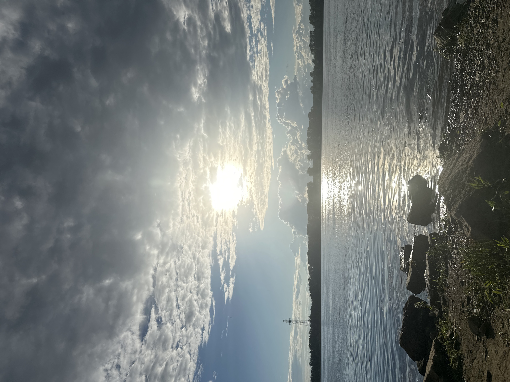 The image shows a wide river, likely the Kama River, in Chaikovsky, Russia, under a dramatic sky filled with a mix of bright sunlight and dark, textured clouds. The sun is partially obscured by clouds, creating a bright, diffused light that reflects strongly on the rippling water's surface. The foreground features a rocky shoreline with scattered rocks and some sparse vegetation. In the middle ground, a dense line of trees forms the opposite bank of the river, and a tall, lattice-style power transmission tower stands out against the horizon on the left side. The water appears calm with gentle waves. The overall scene is natural and serene, with a touch of industrial presence from the power tower.