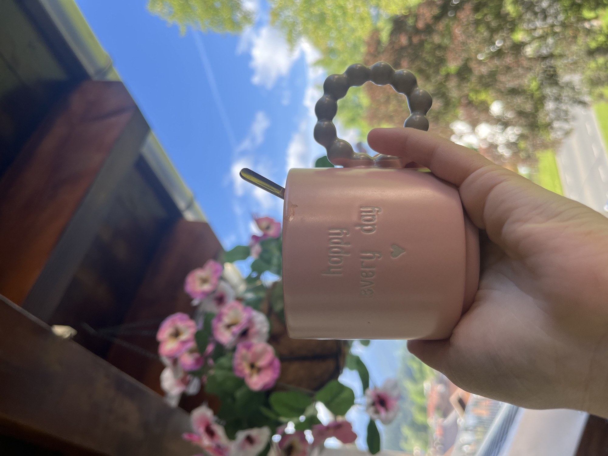 An outdoor shot taken from a slightly low angle shows a hand holding a pink mug with the inscription "happy every day" and a small heart. The mug has a textured, grey handle. A spoon is visible inside the mug. In the background, a hanging basket of pink flowers is visible. Beyond the flowers and mug, a bright blue sky with scattered white clouds is visible, with green trees and hints of buildings in the distance. The overall scene suggests a pleasant, possibly sunny day, with a relaxed atmosphere. The location is the city of Garmisch-Partenkirchen, Germany.
