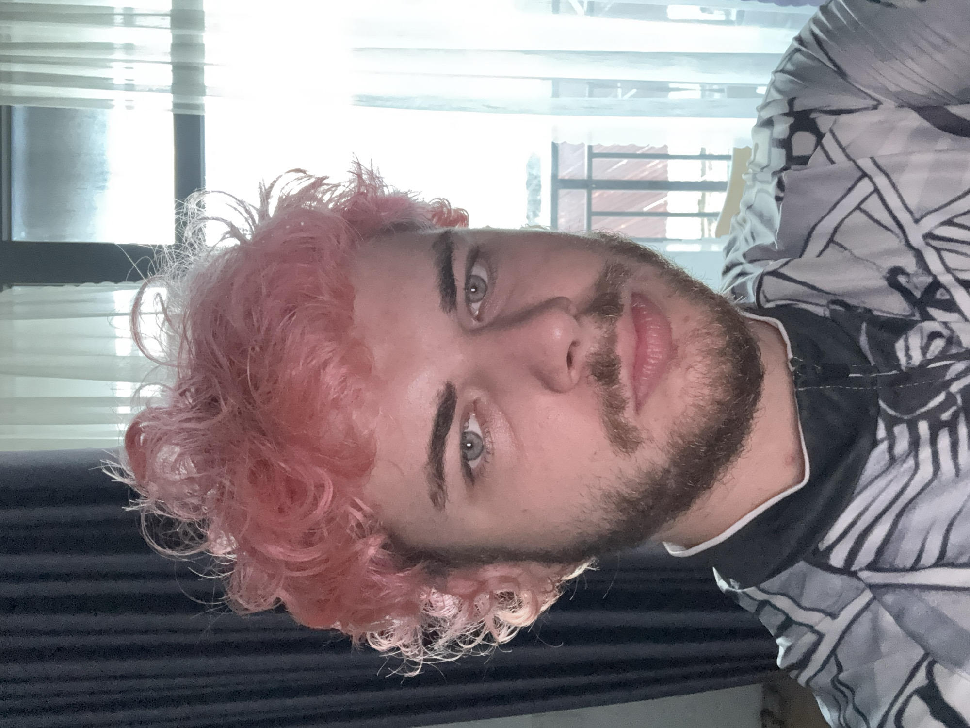 A close-up, portrait-style photograph depicts a man with pink curly hair and a beard. His eyes are light colored. He is wearing a black and white patterned shirt. The background includes a window with white blinds, revealing an exterior cityscape. Based on the architectural style visible in the window, including red-tiled roofs and window frames, the image appears to be taken in Đà Nẵng, Vietnam.