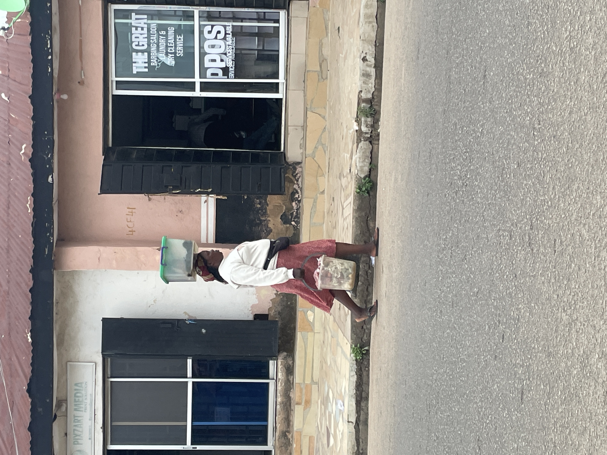 This daytime image captures a street scene in Jos, Nigeria, with a woman walking along a paved sidewalk adjacent to an asphalt road. The woman, likely a street vendor, is dressed in a light-colored long-sleeved top and a reddish-pink skirt, wearing flip-flops and carrying a small black bag across her body. She expertly balances a light green plastic container on her head while holding a clear plastic bucket filled with white items, possibly food or flowers, in her right hand.
The sidewalk is constructed from a mix of light-colored tiles and concrete, with small patches of greenery emerging from cracks. Buildings line the street, indicating a commercial or mixed-use area. On the left, a building features a corrugated reddish-brown roof and a shop with a sign that reads "PIXZART MEDIA PRINT SOLUTION." To the right of the woman, another building with a peach-colored wall displays large glass windows. Signage on these windows identifies businesses as "THE GREAT BARBING SALOON LAUNDRY & DRY CLEANING SERVICE" and "PPOS SERVICES AVAILABLE." A numerical code "4CF41" is also visible on the wall. A person can be vaguely seen inside one of the businesses through the windows. The lighting suggests clear weather.