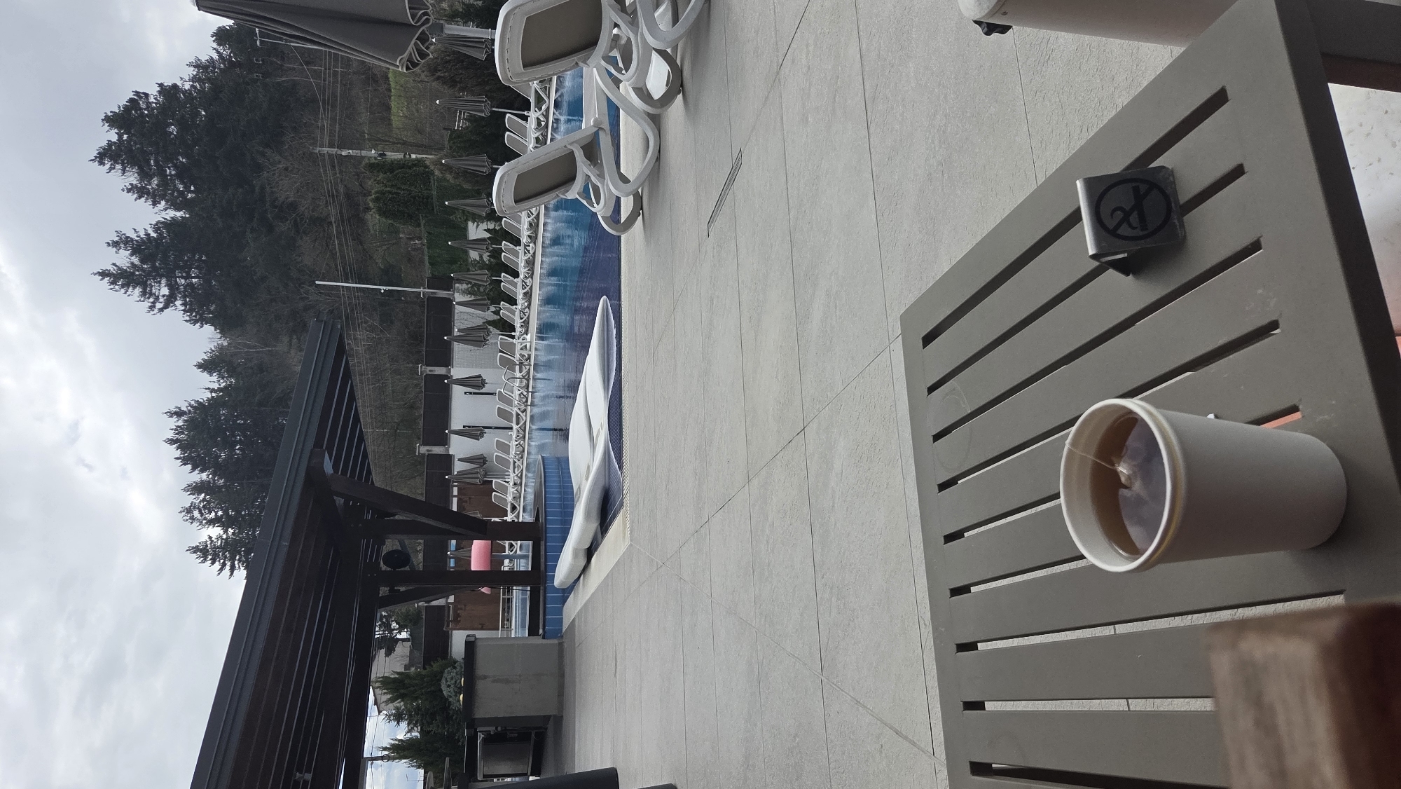 The image captures a poolside scene in Bania, Bulgaria. A gray, slotted patio table is in the foreground, holding a cup of tea and an ashtray. Beyond the table, there's a swimming pool with white lounge chairs lined up along its edge. The pool is part of an outdoor area with a covered structure to the left, providing shade. In the background, a hillside covered with trees is visible, set against a cloudy sky. The scene appears still, with no people immediately visible.