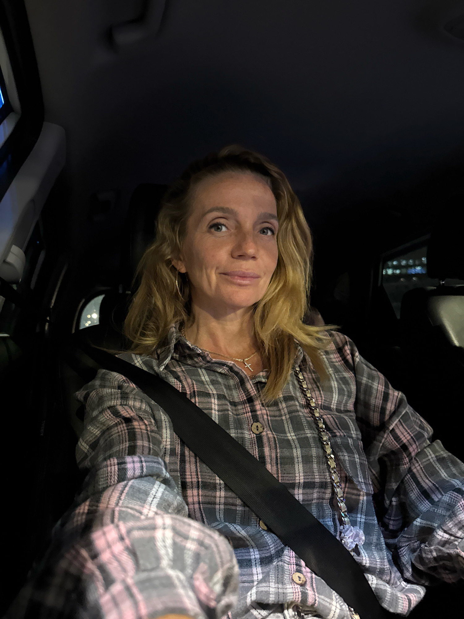 A woman with blonde hair is sitting in the driver's seat of a car at night. She is wearing a plaid shirt and a seatbelt. The interior of the car is dark, with some lights visible from the city outside. The woman is looking up and to the right with a slight smile. Her jewelry includes a necklace with a cross pendant and hoop earrings. The scene appears to be in St. Petersburg, Russia, judging by the dimly lit urban background and the style of the car's interior.