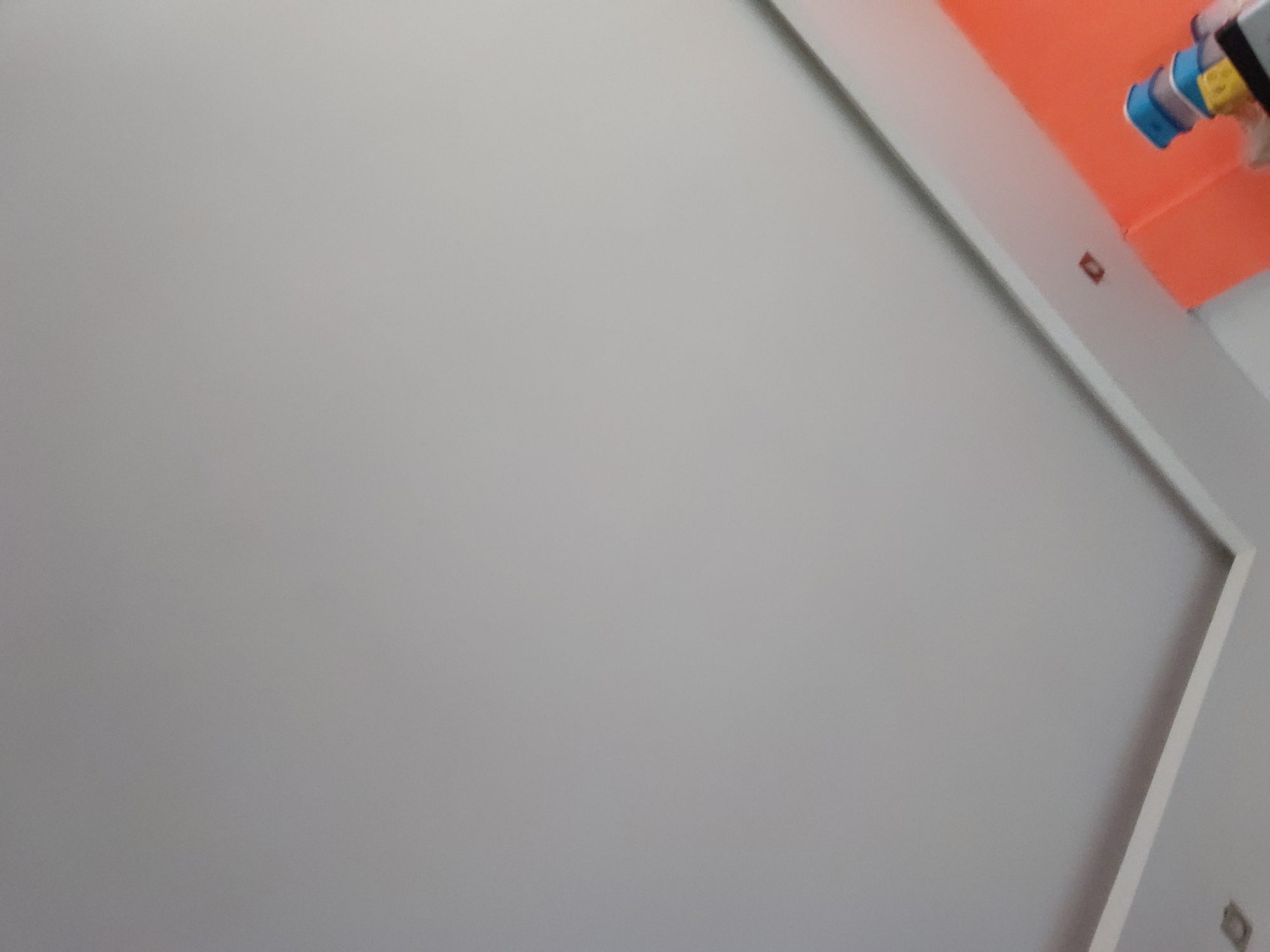 This image captures an interior scene, likely a room undergoing construction or renovation in Santa Ana, Venezuela. The dominant feature is a smooth, light gray wall that fills most of the frame, suggesting it has been freshly painted or prepared.
In the upper right portion of the image, a vibrant orange ceiling contrasts sharply with the gray wall. A distinct, white angular trim or molding runs along the junction between the gray wall and the orange ceiling, creating a clean, modern line. Another section of this white trim is visible along the lower right edge of the gray wall.
Small, reddish-brown rectangular objects, possibly electrical outlet boxes or junction boxes, are visible: one is embedded within the white trim near the top right, and another, less clear, dark rectangular object is located in the bottom right corner of the gray wall, suggesting further electrical points.
In the extreme upper right corner, partially out of focus, is a multi-colored object composed of blue, yellow, and white/gray plastic or ceramic components. Its blurry nature makes its exact function unclear, but it could be part of a fixture, such as a showerhead, a faucet unit, or a ventilation component.
The scene appears static, indicating a moment during or after interior finishing work. There are no people visible, and no discernible activity is taking place. The lighting is even, but the time of day and external weather conditions cannot be determined from this indoor perspective. No text is visible in the image. The combination of the gray wall, orange ceiling, and white angular trim points to a contemporary design aesthetic.