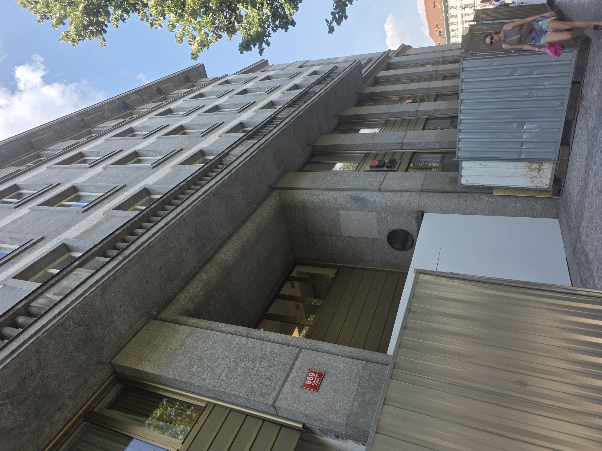In an exterior, low-angle, medium shot, a large, grey stone building with many windows dominates the frame, extending upwards into a bright blue sky with scattered clouds. The building features a protruding overhang supported by columns, with a balustrade running along its edge. A red street sign with the number "969" is affixed to the lower left portion of the building's facade. In the foreground, temporary metal and white barriers are erected along the sidewalk, suggesting construction or renovation work. A young woman wearing a tank top, denim shorts, sunglasses, and a cross-body bag, holding a pink bag, walks away from the camera to the right of the frame. Further down the street, other buildings with terracotta roofs are visible in the distance. A large tree with green foliage branches into the upper right of the image. The overall scene depicts an urban environment on a sunny day.