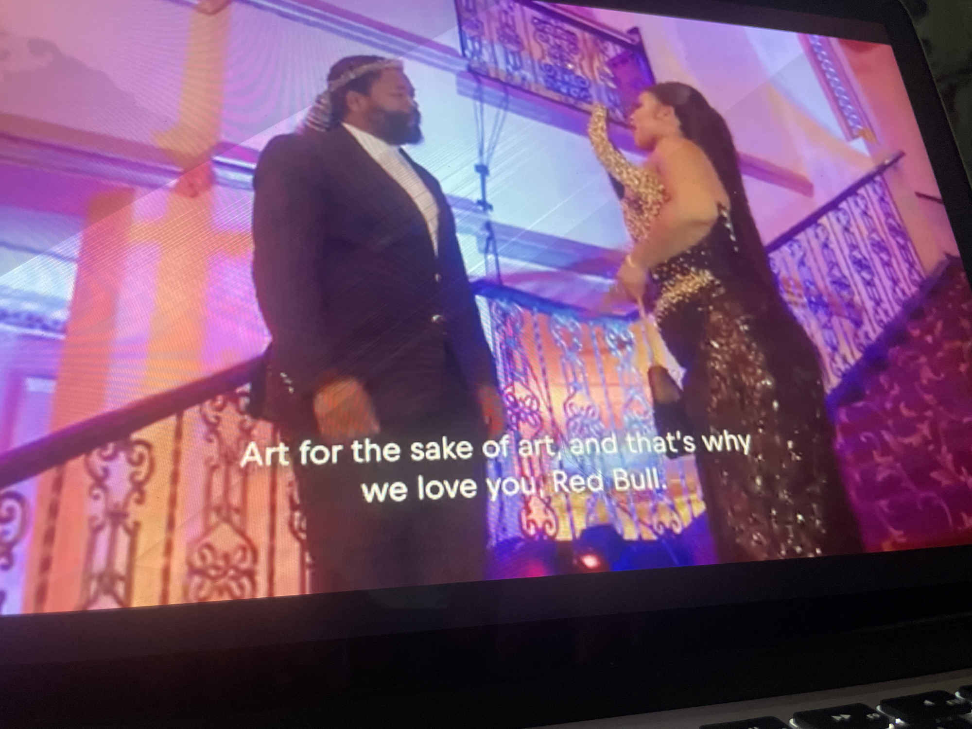 The image shows a laptop screen displaying a video with subtitles. Two people are visible on the screen. A man on the left, wearing a black suit and a white shirt with a patterned tie, has a beard and is wearing a headscarf. A woman on the right, wearing a sequined dress, is reaching her arm up. The background appears to be a staircase with decorative railings, illuminated by colorful lights. The subtitles read: "Art for the sake of art, and that's why we love you, Red Bull." The scene takes place indoors, possibly at an event. The exact location within the city of Unknown, Unknown is not determinable from the image.