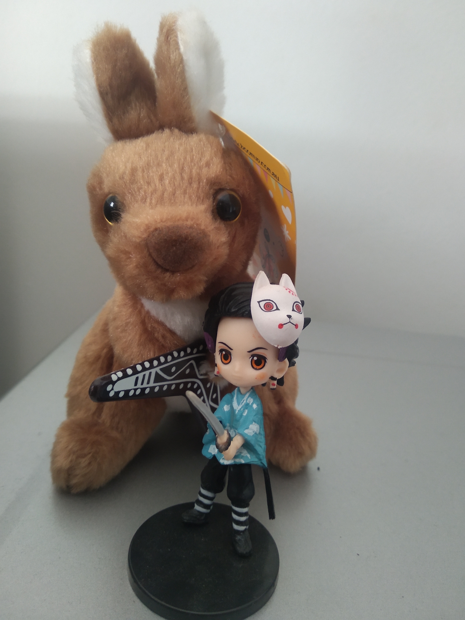 An indoor close-up photo depicts a stuffed rabbit toy and a small anime figurine. The plush toy is brown with white inner ears and large black eyes. It is positioned behind the figurine and appears to be holding a black object with white markings, resembling a stylized chain. The anime figurine is a young boy with dark hair, large orange eyes, and a white fox mask with red markings on his face. He is wearing a light blue patterned kimono and black pants with white striped socks. The figurine stands on a black circular base. The background is a plain, light gray wall. The lighting is even, suggesting an indoor setting with artificial light. The scene is arranged on a gray surface, possibly a table or shelf. There is no visible indication of the specific location within South Tangerang, Indonesia.