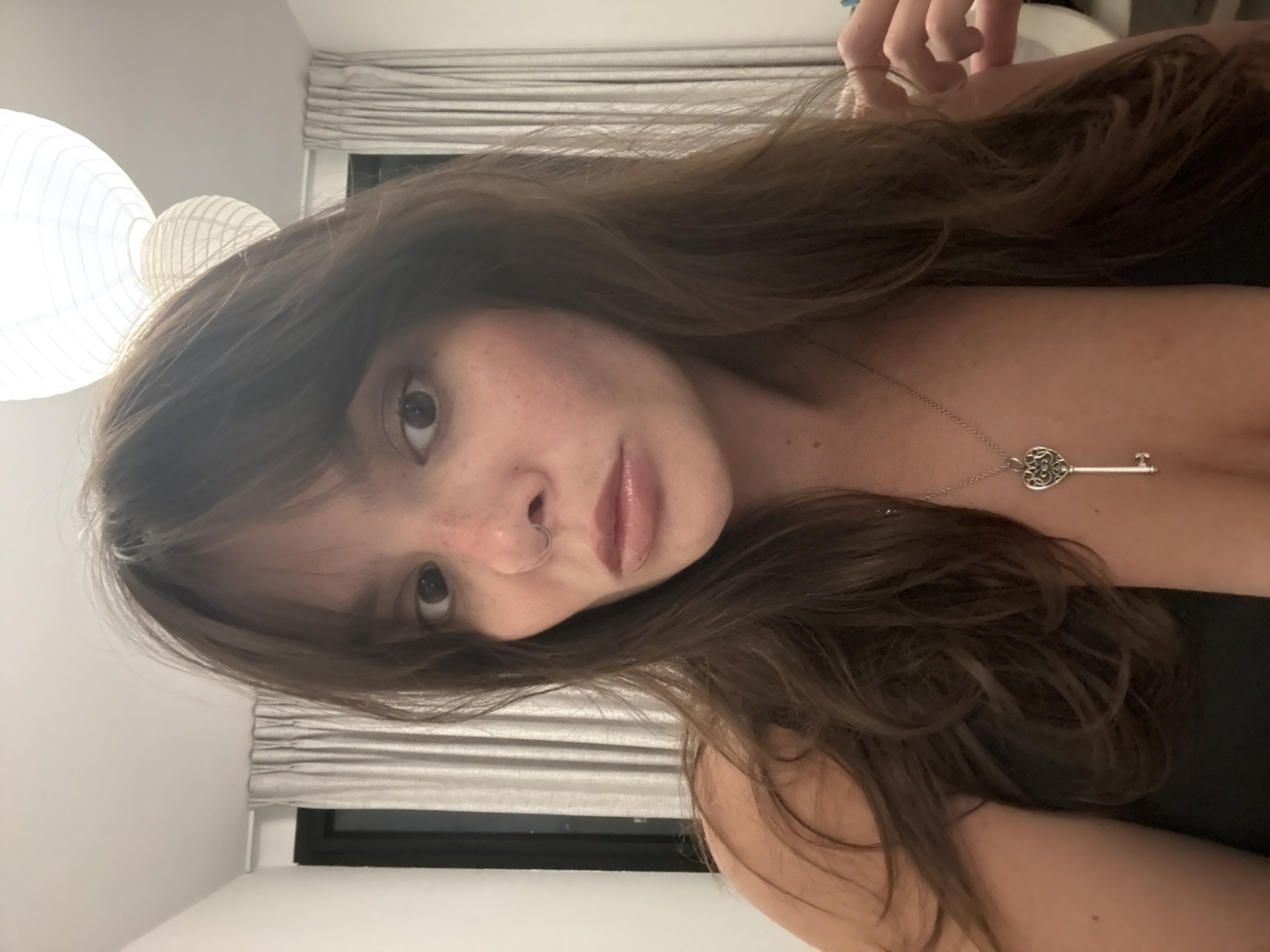 A woman with brown hair and freckles is looking into the camera. She is wearing a necklace with a key pendant. The background is blurred and shows curtains and lamps. The image was taken indoors. The image does not contain any information about the location in the city of Tel-Aviv, Israel.