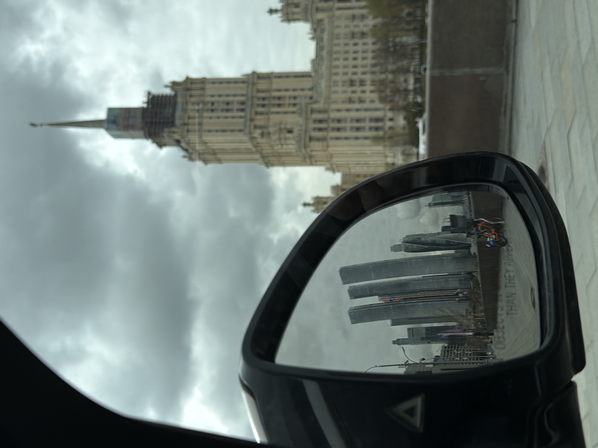 The image captures a scene in Moscow, Russia, featuring the Kotelnicheskaya Embankment Building in the background under a cloudy sky. A car's side mirror reflects the Moscow International Business Center skyscrapers. A pedestrian with a bicycle is also visible in the mirror. The mirror bears the text "OBJECTS IN MIRROR ARE CLOSER THAN THEY APPEAR". The car interior is partly visible on the left side of the image. The foreground is composed of a paved surface.