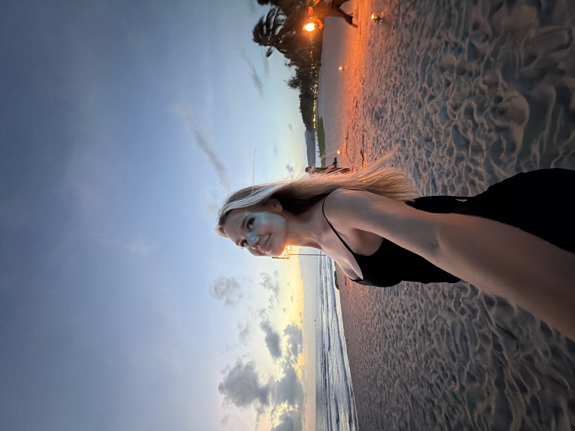 A woman is standing on a sandy beach at dusk, looking up towards the sky. She is wearing a black sleeveless dress. The sky is a gradient of blue and orange, with some clouds. The ocean is visible to her left, with gentle waves rolling onto the shore. In the background, there are palm trees and lights from a resort or buildings. There are several small, lit torches or lanterns on the beach, and a person can be seen in the distance holding a torch. The scene appears to be on a tropical beach in Choeng Thale, Thailand, likely during a fire performance or similar evening event.