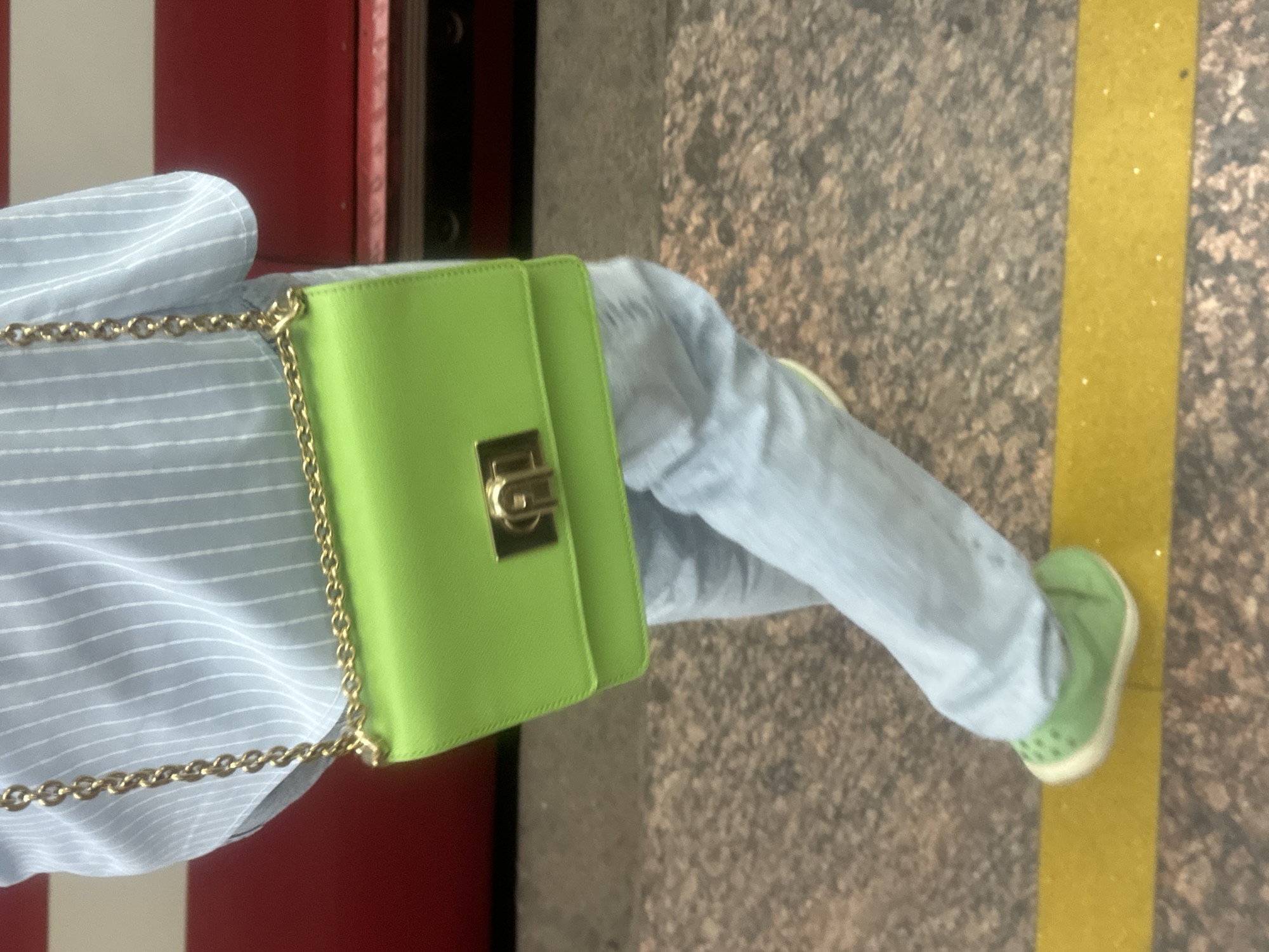 This image captures a partial view of an individual walking on a subway platform in Saint Petersburg, Russia. The individual is seen from approximately the mid-torso downwards, wearing a light blue shirt with thin vertical white stripes, and light blue trousers or jeans. A bright lime green rectangular shoulder bag with a gold chain strap and a prominent gold rectangular clasp is carried on the individual's right side. The individual's feet are clad in bright lime green slip-on shoes, possibly with perforations on the upper and a light-colored sole. The footwear is positioned over a wide, bright yellow safety line on the platform floor. The platform itself features a speckled, possibly granite, surface in shades of grey and brown. In the background, a red subway train car is partially visible, with a darker lower section and a lighter, possibly beige or white, strip above the red. The scene suggests movement, with the individual in mid-stride.