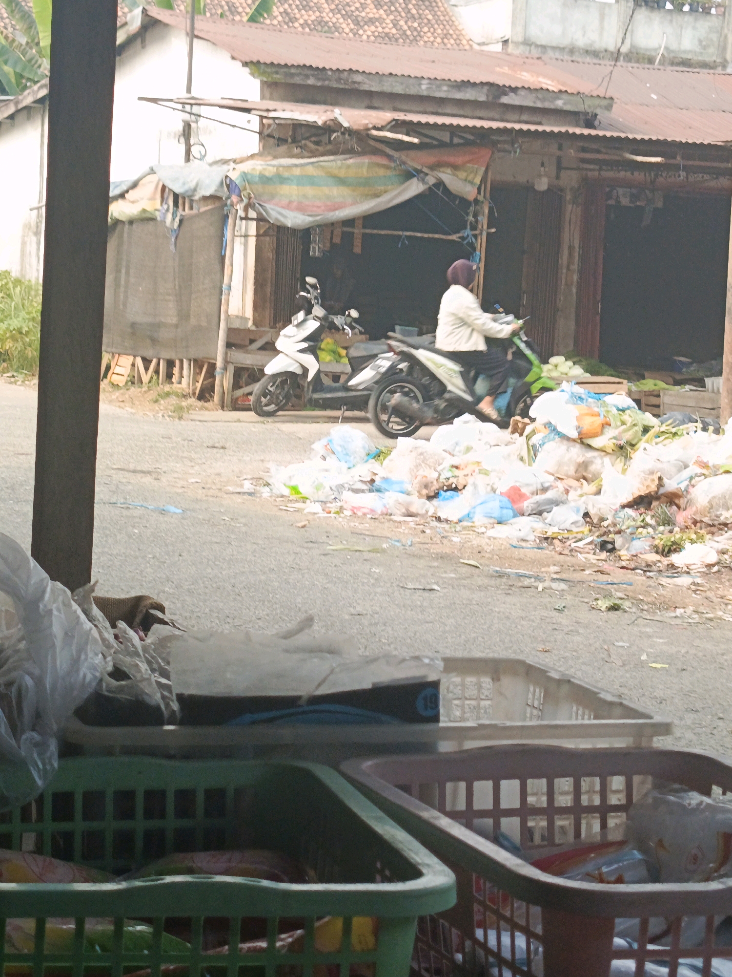 In the city of Megang Sakti I, Indonesia, a scene of outdoor commerce and refuse is depicted. A white scooter is parked in front of a rustic, open-fronted structure with a corrugated metal roof. A person wearing a light-colored jacket and a headscarf is seated on the scooter, facing left. To the right of the scooter, a large pile of garbage, composed of various plastic bags, wrappers, and organic waste, is accumulated on the asphalt ground. In the foreground, out of focus, are two plastic baskets, one green and one brown, containing items that are not clearly discernible. The background reveals the side of a building with a white wall and more corrugated metal roofing. The overall environment suggests a market or roadside stall area with considerable litter.