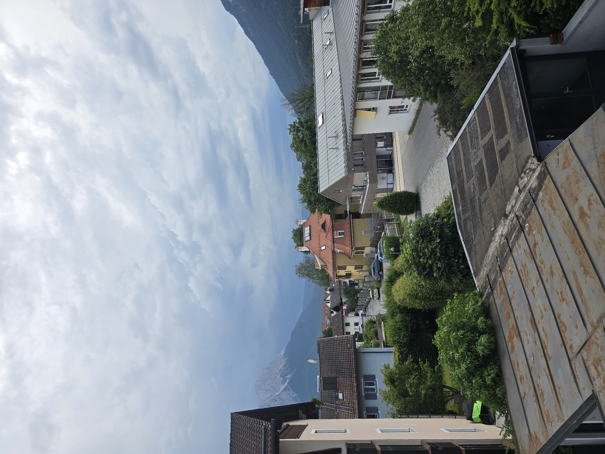 This is a landscape photograph taken in Garmisch-Partenkirchen, Germany, with the camera oriented vertically. The scene depicts a residential area nestled against a backdrop of lush green mountains under a partly cloudy sky. In the foreground, a weathered, corrugated metal roof is visible, suggesting a rooftop perspective.
The midground showcases a street lined with multi-story buildings, characterized by their pitched roofs, balconies, and numerous windows. These structures exhibit a variety of architectural styles and colors, with some featuring warm yellow facades and others a more modern, gray metal roof. Dense green trees and foliage are interspersed throughout the residential area, adding to the natural beauty of the setting.
In the distance, the imposing presence of a large, verdant mountain range dominates the horizon. The sky is filled with a mix of white, wispy clouds and darker, more textured clouds, hinting at a dynamic weather system, possibly during the day. The overall impression is one of a tranquil, scenic town in a mountainous region. No people are clearly visible in the image, and no text is discernible.