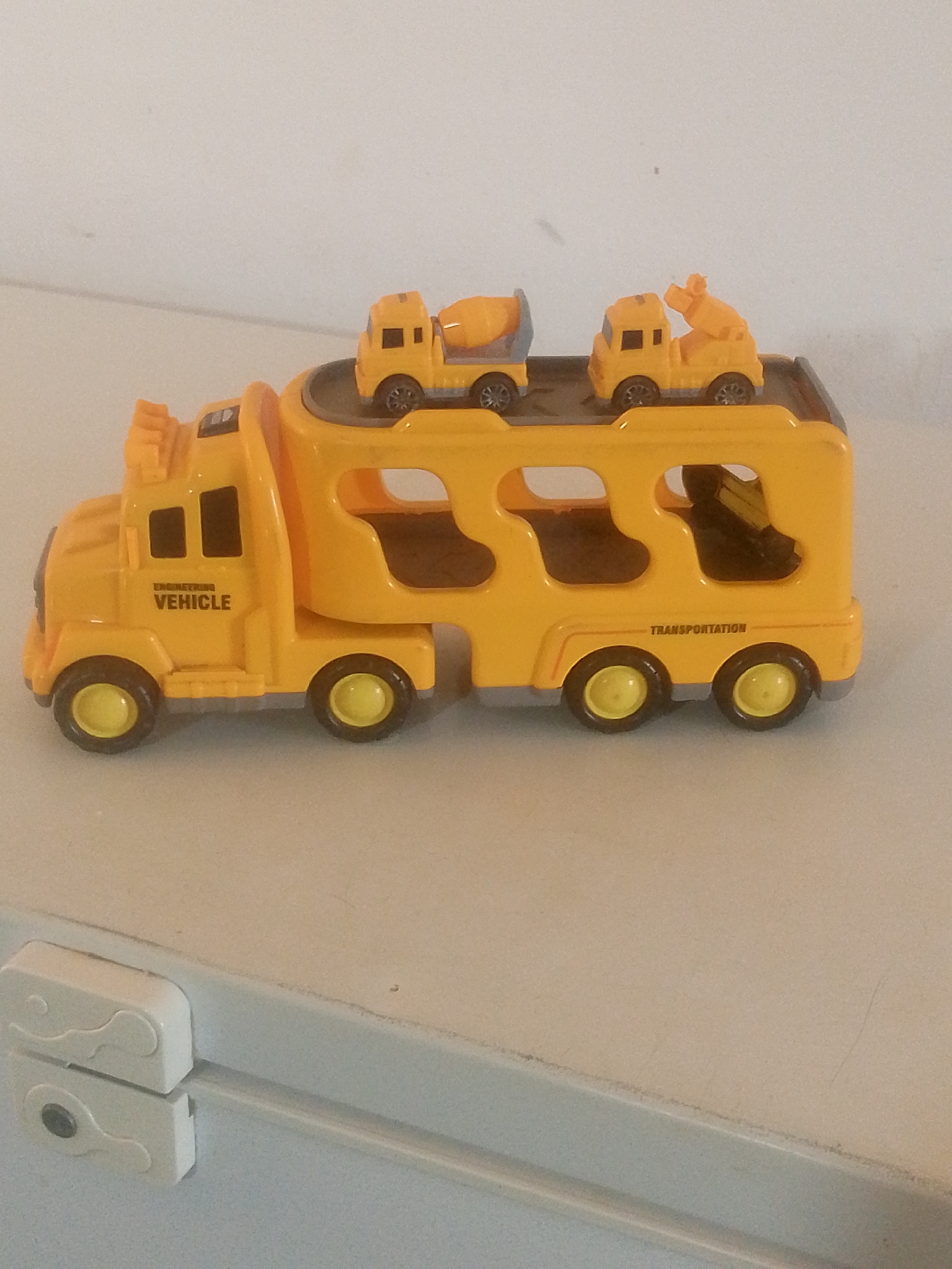 A toy truck carrier is positioned on a light-colored surface, possibly a table or shelf, in front of a white wall. The carrier is bright yellow and has two levels. The lower level has cutouts, and the upper level is a platform. Two smaller toy trucks, one resembling a cement mixer and the other a tow truck, are placed on the upper platform of the carrier. The toy truck carrier has the word "VEHICLE" printed on its side. The background appears to be a plain white wall. The lighting suggests an indoor setting. The image does not contain any specific details that would indicate its location in La Tagua, Venezuela.