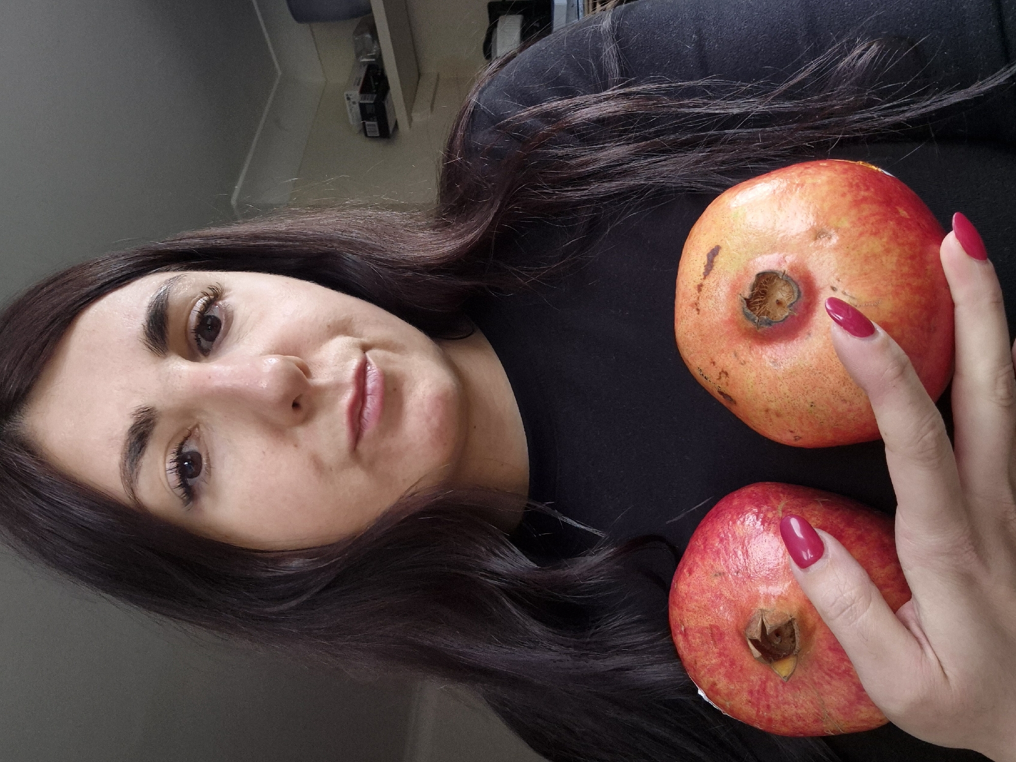 A brunette woman with fair skin is photographed indoors, lying on a dark-colored surface, possibly in West Suffolk, United Kingdom. Her hair is spread out around her, and she is looking toward the camera. Two pomegranates are positioned near her face and hair. Her right hand, with red fingernails, is near one of the pomegranates. The background is indistinct, suggesting an indoor setting near a wall and possibly some shelves.