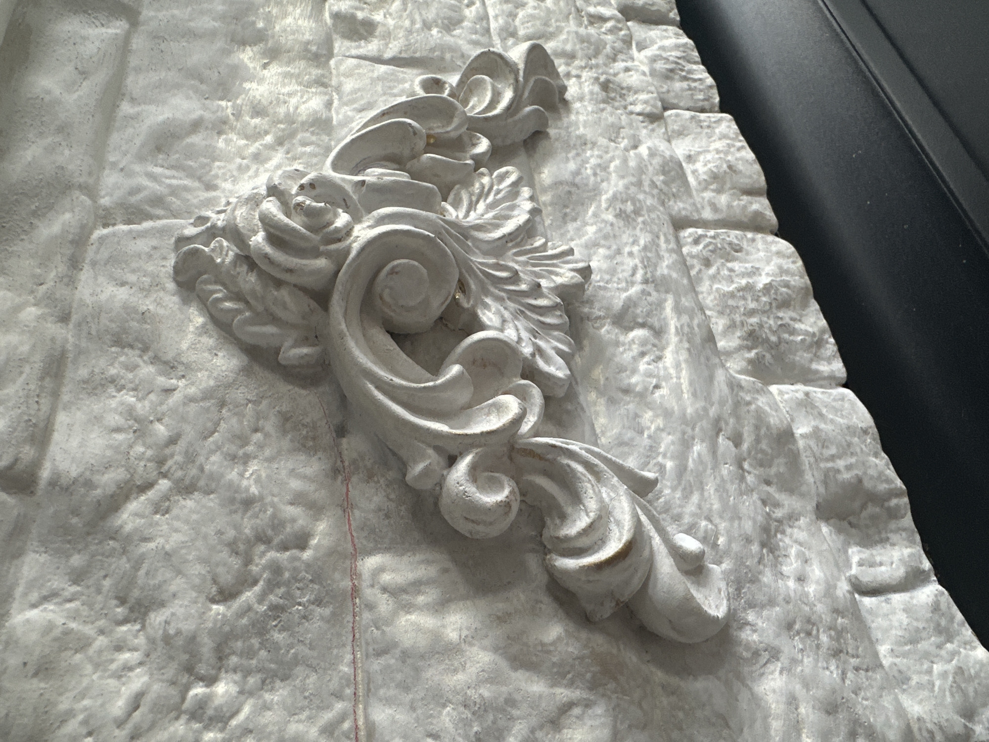 A white ornate carving is affixed to a white stone-like wall. The carving is in a rococo style, featuring scrolling acanthus leaves and a rose motif. A thin red thread is visible across the wall to the left of the carving. The wall appears to be textured and possibly artificial, resembling rough-hewn stones. A dark surface, likely a mantelpiece or shelf, is visible in the lower right corner, casting a shadow on the wall. The lighting suggests an indoor environment with a light source to the right.