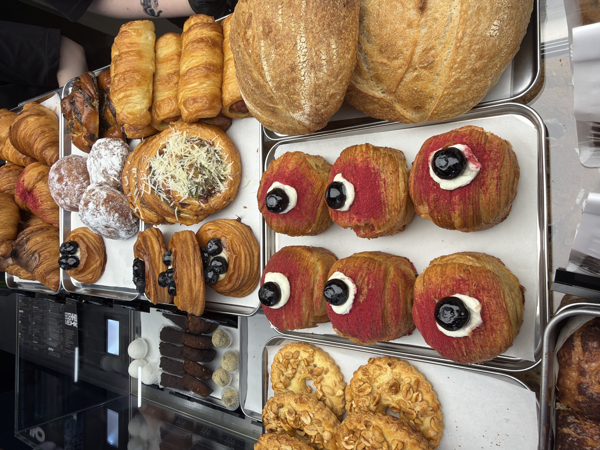 A high-angle, close-up shot depicts a selection of pastries arranged on metal trays in a display case, possibly in a bakery. Two large loaves of bread occupy the top right. Below them are six round pastries covered in red sprinkles and topped with white icing and a black cherry. Several pastries filled with blueberries are to their left, as well as a larger pastry with white cheese topping. To the left of those are powder-covered pastries and plain croissants. To the left of all the pastries is a container holding white balls, chocolate sticks, and green balls. In the lower center of the image are golden, ring-shaped pastries with peanut pieces. The name "Ижевск" is visible on the menu in Cyrillic, indicating the location as Izhevsk, Russia. A person's arm with tattoos is visible in the upper left corner.