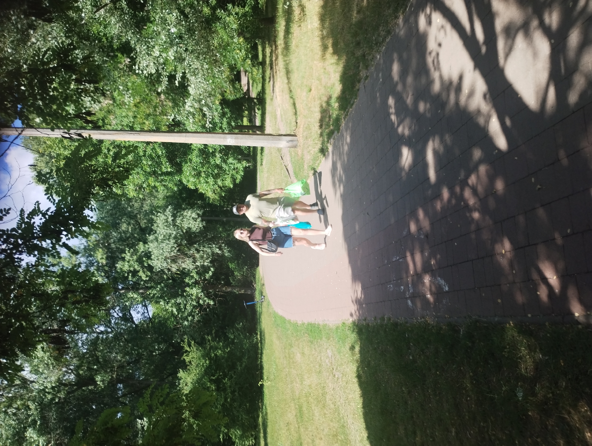 Two individuals are walking on a paved path through a park. A woman, wearing a dark brown tank top and denim shorts, walks ahead of a man wearing a light green t-shirt, grey shorts, and a baseball cap. Both are carrying plastic bags. The path is lined with grass and trees, and a lamppost stands to the right of the path. The scene appears to be illuminated by sunlight, casting shadows on the ground.