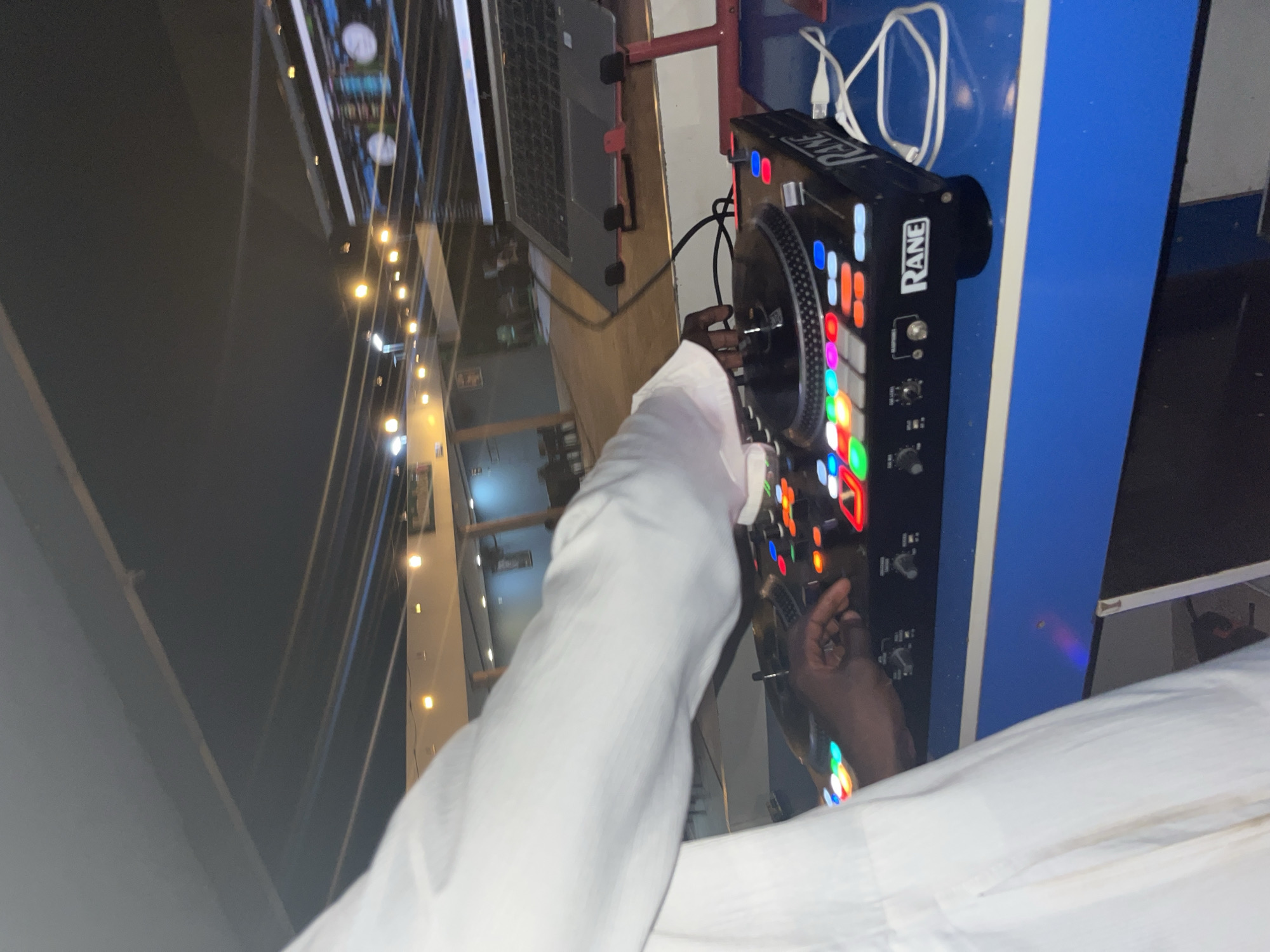 In an outdoor, nighttime setting, a person wearing a white robe is operating a DJ turntable and mixer. The DJ equipment, labeled "RANE," features numerous illuminated buttons. A laptop is visible on a stand to the right of the DJ setup. The background shows string lights, a building, and tables with chairs, suggesting a venue or event space. The ground appears to be paved.