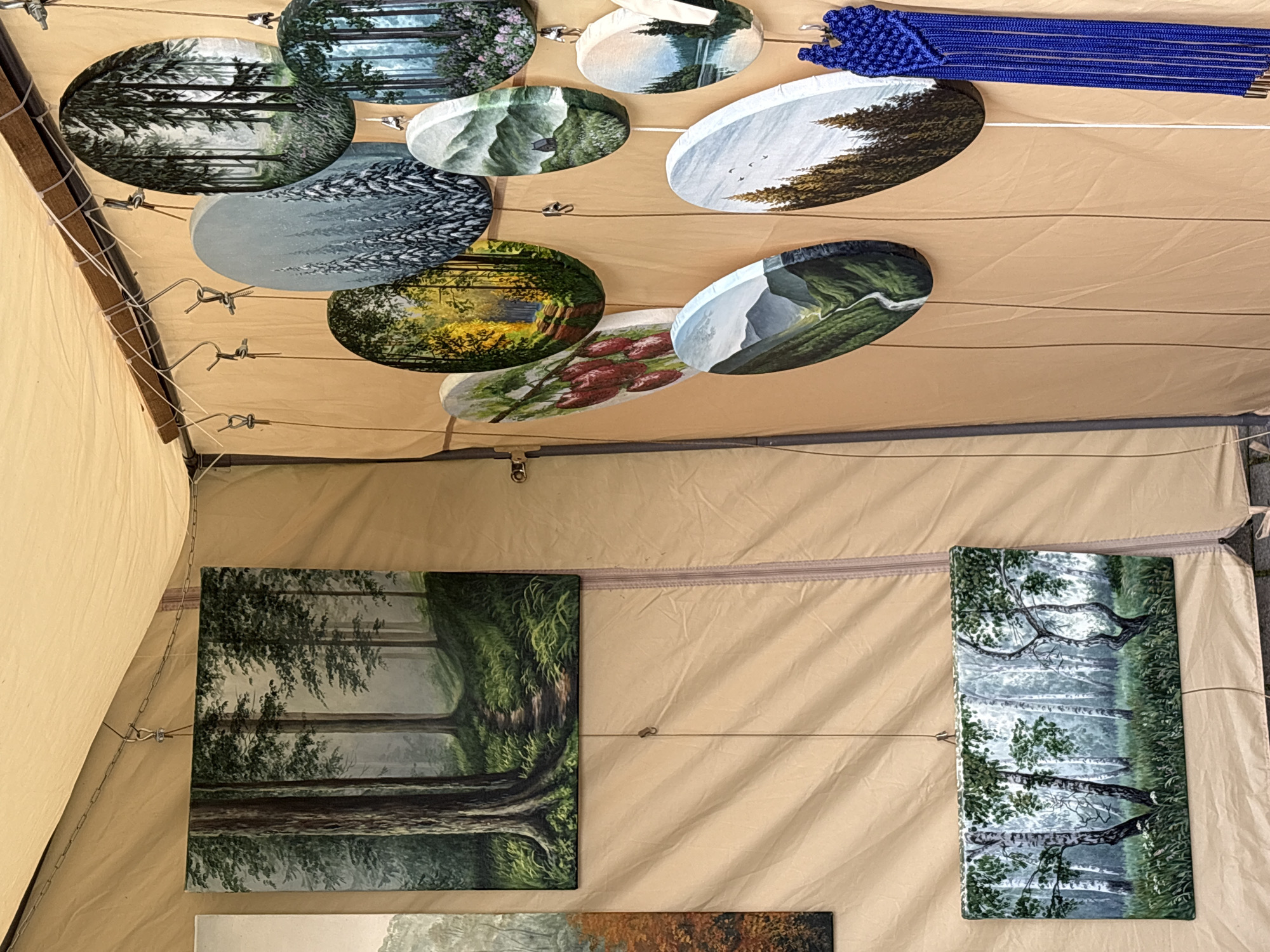 The image shows a display of paintings, likely at an outdoor market or exhibition, under a tan-colored canopy. Several rectangular and oval paintings, primarily featuring nature scenes such as forests, mountains, and a lake, are hung on a wire. One large rectangular painting on the left depicts a forest path with tall trees. Below it, another rectangular painting shows a birch forest. To the right, a collection of oval paintings showcases various landscapes, including a snowy forest, a sunlit forest path, a mountain valley, and a body of water with trees. A blue macrame decoration hangs on the right side of the display. The background consists of the tan fabric of the canopy and supporting structures. No people are visible in the image, and there is no discernible information about the specific location within Vitsebsk, Belarus.