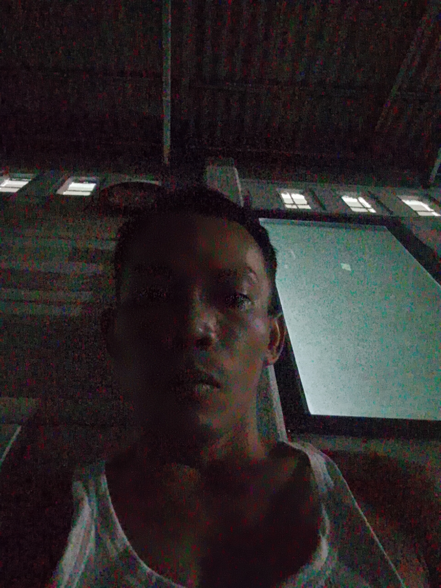 The photograph depicts a person, likely indoors, in low lighting conditions. The person appears to be wearing a light-colored tank top. Above the person are a series of rectangular lights, possibly fluorescent, positioned in a row. To the person's right is a large, bright rectangular object. The location is potentially Bukit Ketok, Indonesia, based on the prompt.