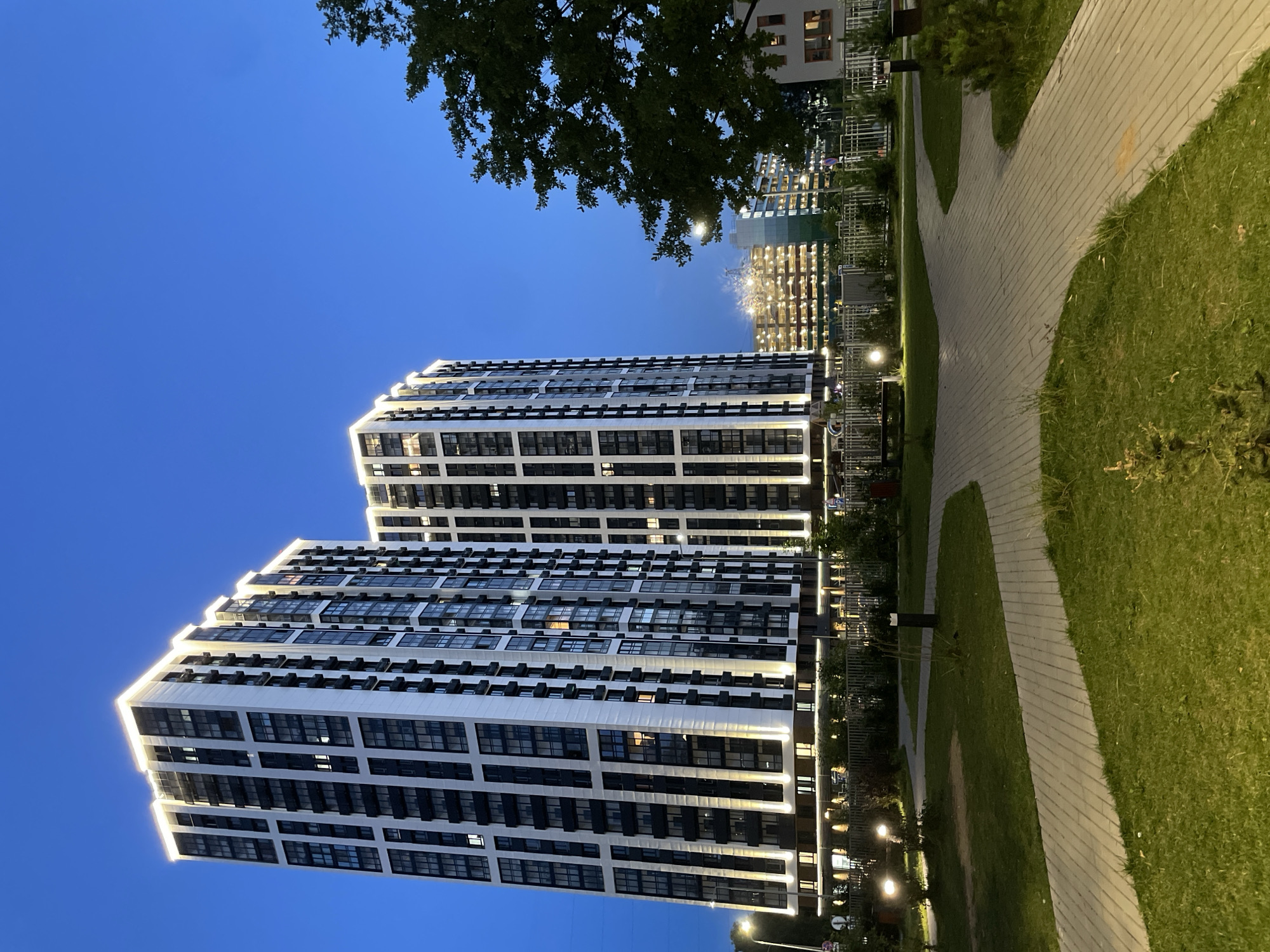 The image captures a modern residential complex in Odintsovo, Russia, at dusk. Two tall, multi-story buildings with a contemporary black and white facade dominate the scene, illuminated by accent lighting along their upper edges and at their bases. The buildings feature numerous windows, some of which are lit, indicating occupancy. In the foreground, a park-like area with manicured lawns and paved walkways is visible. Several lampposts are strategically placed along the paths, casting a warm glow. Beyond the main buildings, other structures, including what appears to be a parking garage or a lower residential building, are also illuminated. A large tree with dense foliage is positioned on the right side of the frame, partially obscuring the sky. The sky is a deep blue, characteristic of twilight. There are no people or vehicles clearly visible in the immediate vicinity of the residential buildings or the park. The overall impression is one of a well-maintained, modern urban environment.