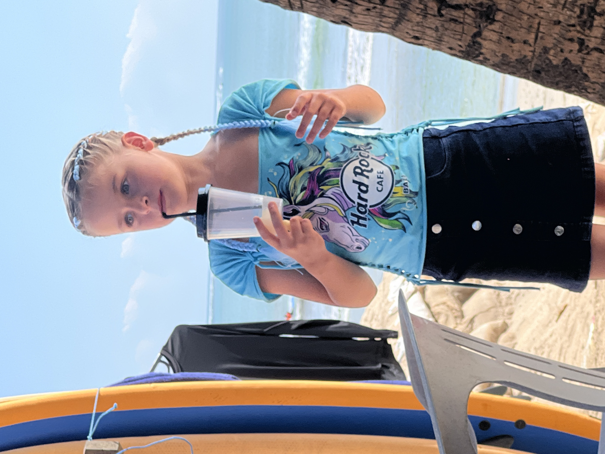 A young Caucasian girl with blonde hair braided with light blue accents stands on a sandy beach, holding a clear plastic cup with a black straw to her lips. She wears a light blue off-the-shoulder top featuring a colorful unicorn graphic and the "Hard Rock Cafe Bali" logo, paired with a dark blue denim skirt with silver buttons. The background shows the ocean with gentle waves under a clear blue sky. To the left, two surfboards, one orange and one blue, are propped up, with a dark garment hanging on a rack behind them. A portion of a gray chair is visible in the foreground, and a palm tree trunk is on the right side of the frame. The scene suggests a casual beachside location, likely in Kuta, Indonesia, given the "Hard Rock Cafe Bali" branding.