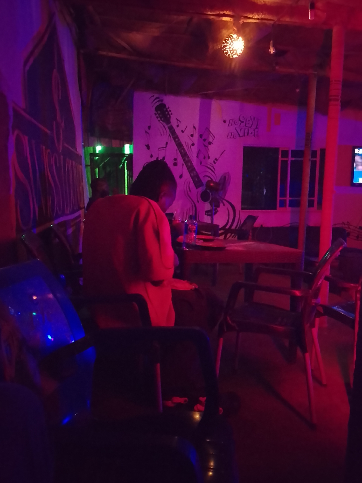 This image captures an indoor scene, likely a bar, lounge, or casual restaurant, characterized by dim, atmospheric lighting. The dominant colors are purple and red, with hints of green and blue from various light sources. The time of day appears to be evening or night.
In the foreground, a person is seated with their back to the viewer, wearing an orange or red top, at a dark table. A plastic water bottle and other indistinct items are on the table. The furniture consists of dark tables and chairs scattered throughout the space.
A notable feature is a large white mural on the back wall, depicting the outline of a guitar surrounded by musical notes. Below the guitar, a small fan is visible on a table. Above the guitar, the text "No SEX! No VIBE!" is clearly written on the mural. A spherical, intricately cut light fixture hangs from the rustic, thatched-like ceiling, casting a warm glow.
On the far left, a large banner or sign is partially visible, displaying text that appears to be "SWAHILISH" or a similar word. In the background to the right, a television screen with a blue glow can be faintly seen. A doorway with a bright green light is visible towards the left background. The overall ambiance is relaxed and casual, suggesting a place for socializing or entertainment.
