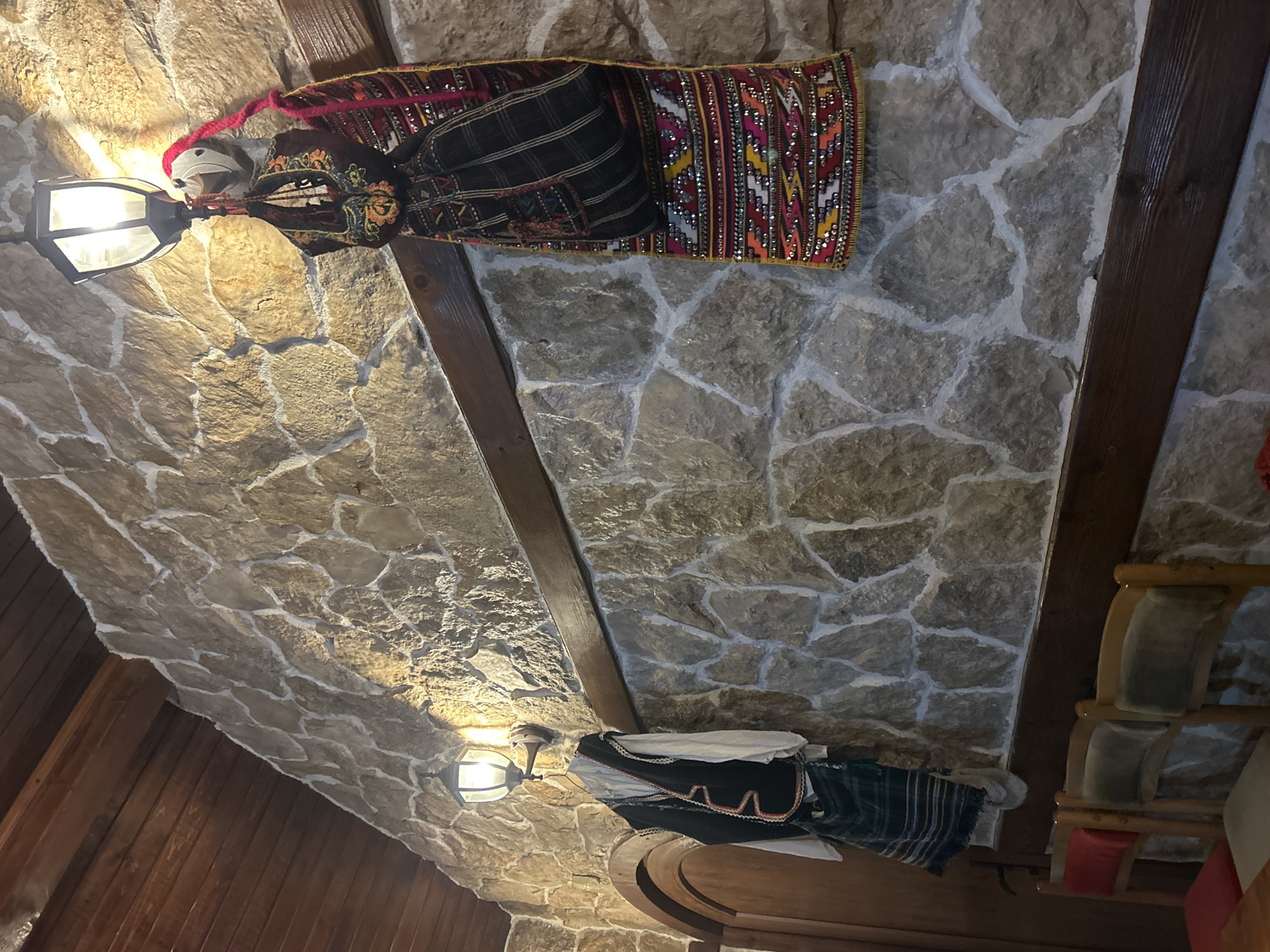 The image shows the interior of a building with stone walls and a wooden-beamed ceiling. On the walls are displayed traditional Bulgarian costumes. One costume is predominantly black with colorful embroidered details and a mirrored pattern, while the other is white, black and green plaid with red and white trim. Two lanterns hang on the wall illuminating the space. The building appears to be located in Mirovyane, Bulgaria, given the traditional Bulgarian cultural items present.